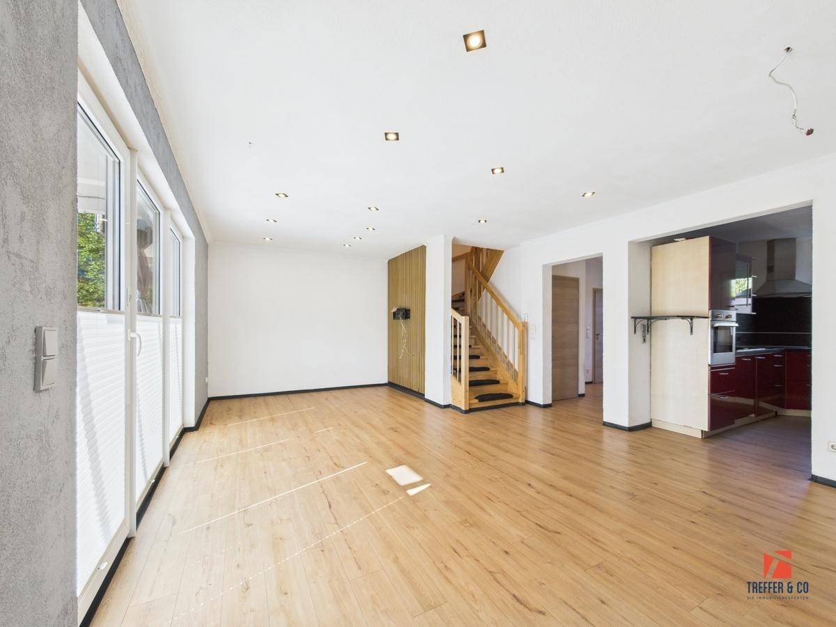 Geräumiges Wohnzimmer mit hellem Laminatboden, großen Fenstern und Zugang zur Küche.