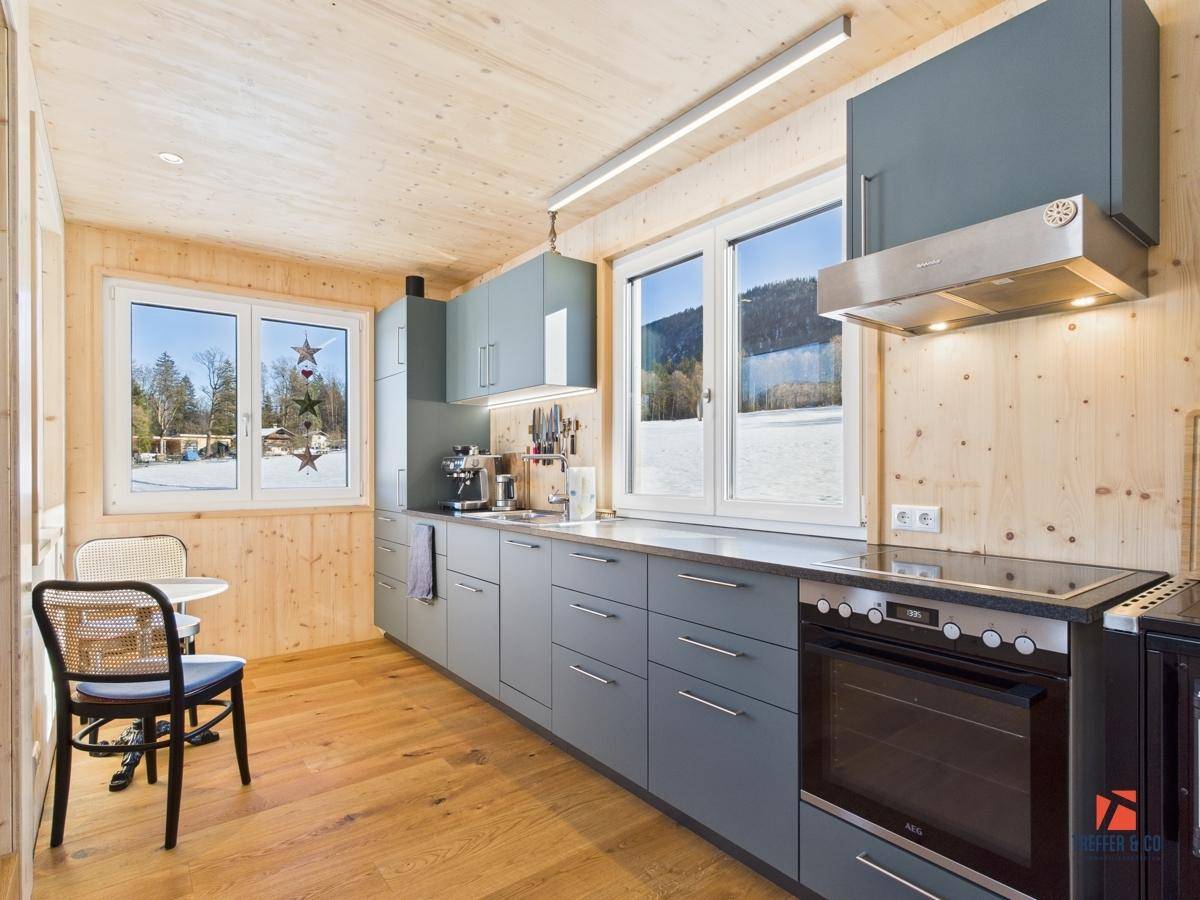 Helle Küche mit Holzverkleidung, blaugrauen Schränken und Blick auf die verschneite Landschaft und einen See.