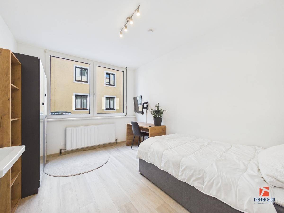 Einfaches Schlafzimmer mit Einzelbett, Schreibtisch und einem großen Fenster, das viel Tageslicht hereinlässt.