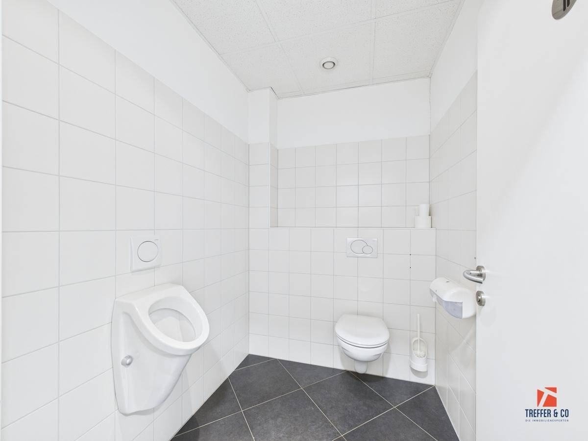 Saubere Herrentoilette mit Urinal, Wand-WC und weißen Fliesen an den Wänden.