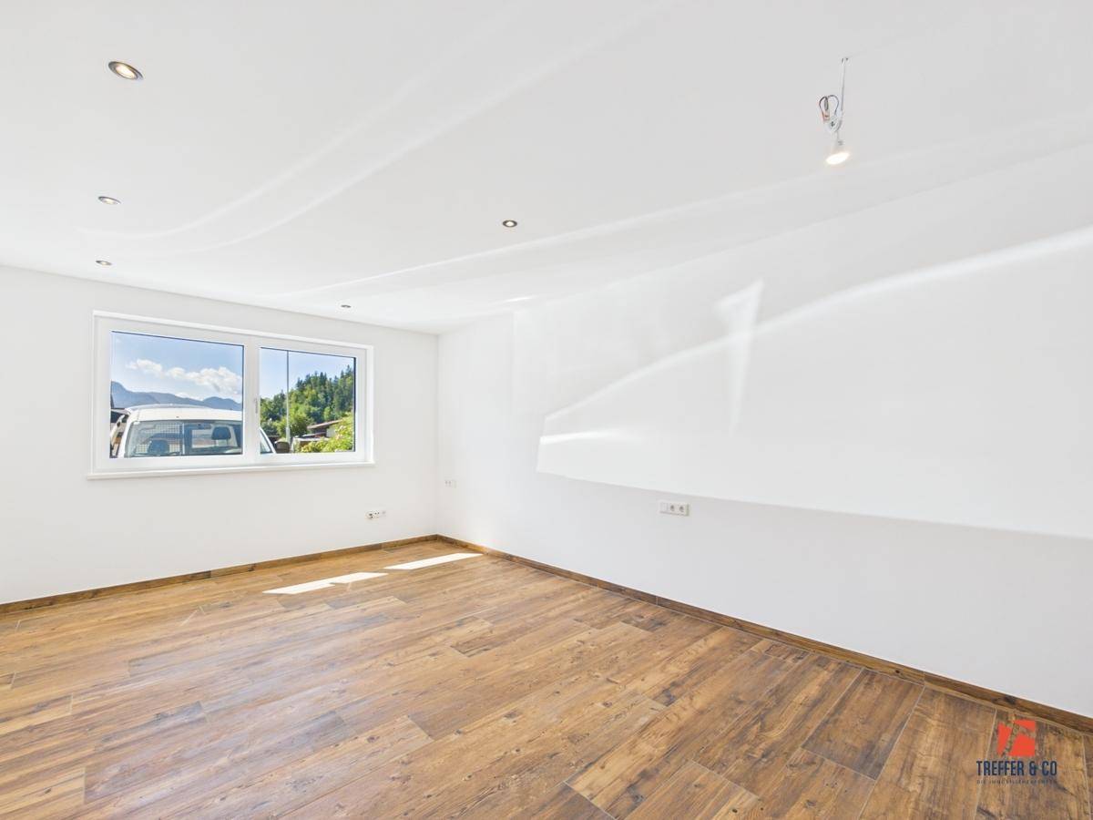 Helles Zimmer mit Holzoptik-Boden und Fensterblick auf die umliegende Landschaft und Berge.