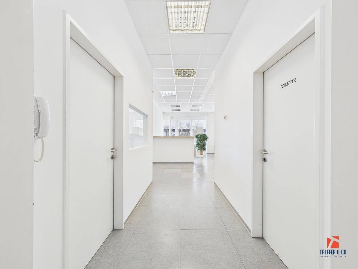 Heller Bürogang mit weißen Wänden, Fliesenboden und Blick auf den Empfangsbereich im Hintergrund.
