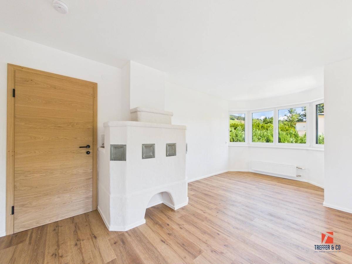 Helles Wohnzimmer mit Holzboden und einem traditionellen Kachelofen, der Gemütlichkeit ausstrahlt.