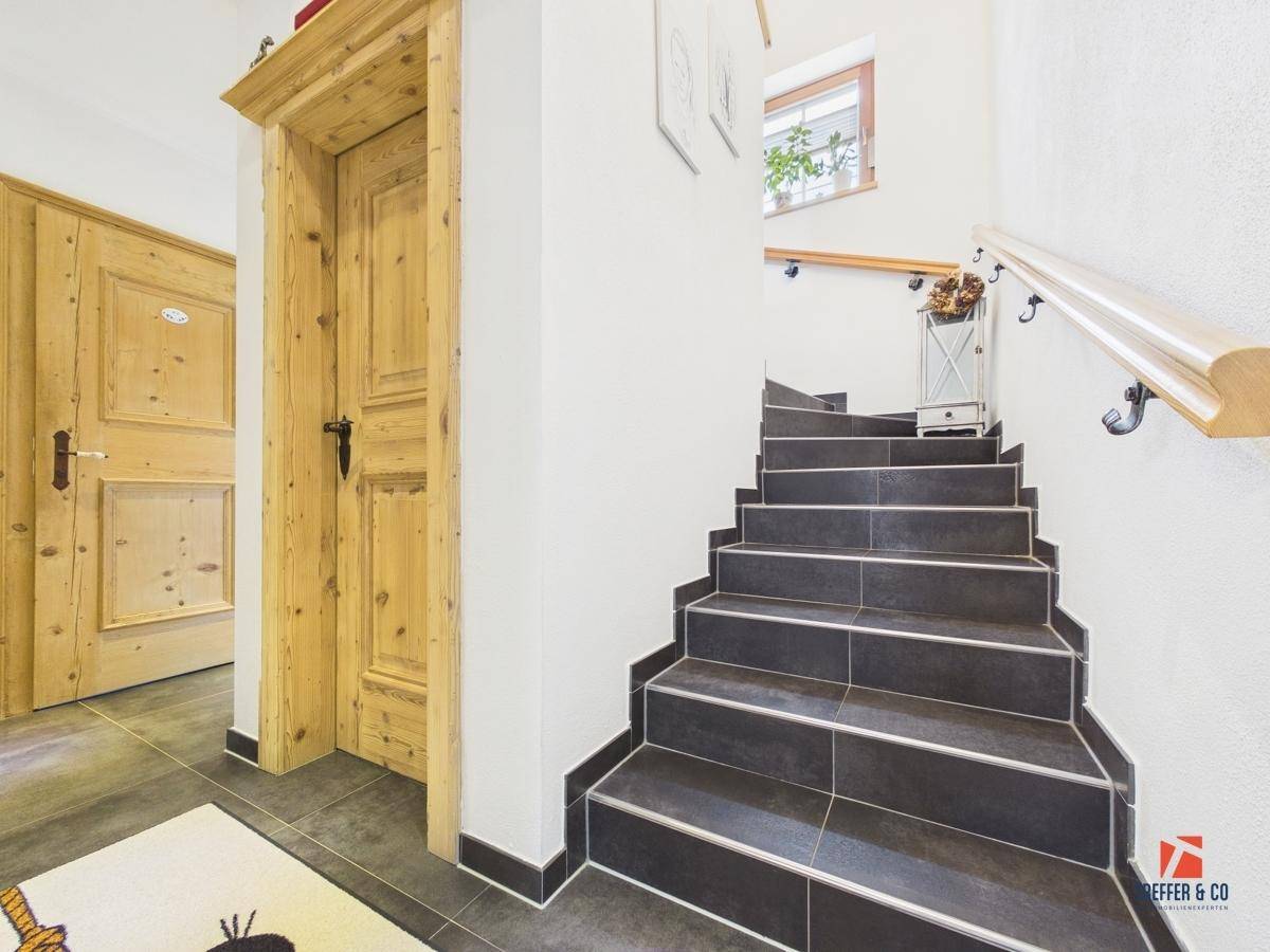 Treppenhaus mit dunklen Fliesenstufen, Holzgeländer und einer Holztür im Landhausstil.