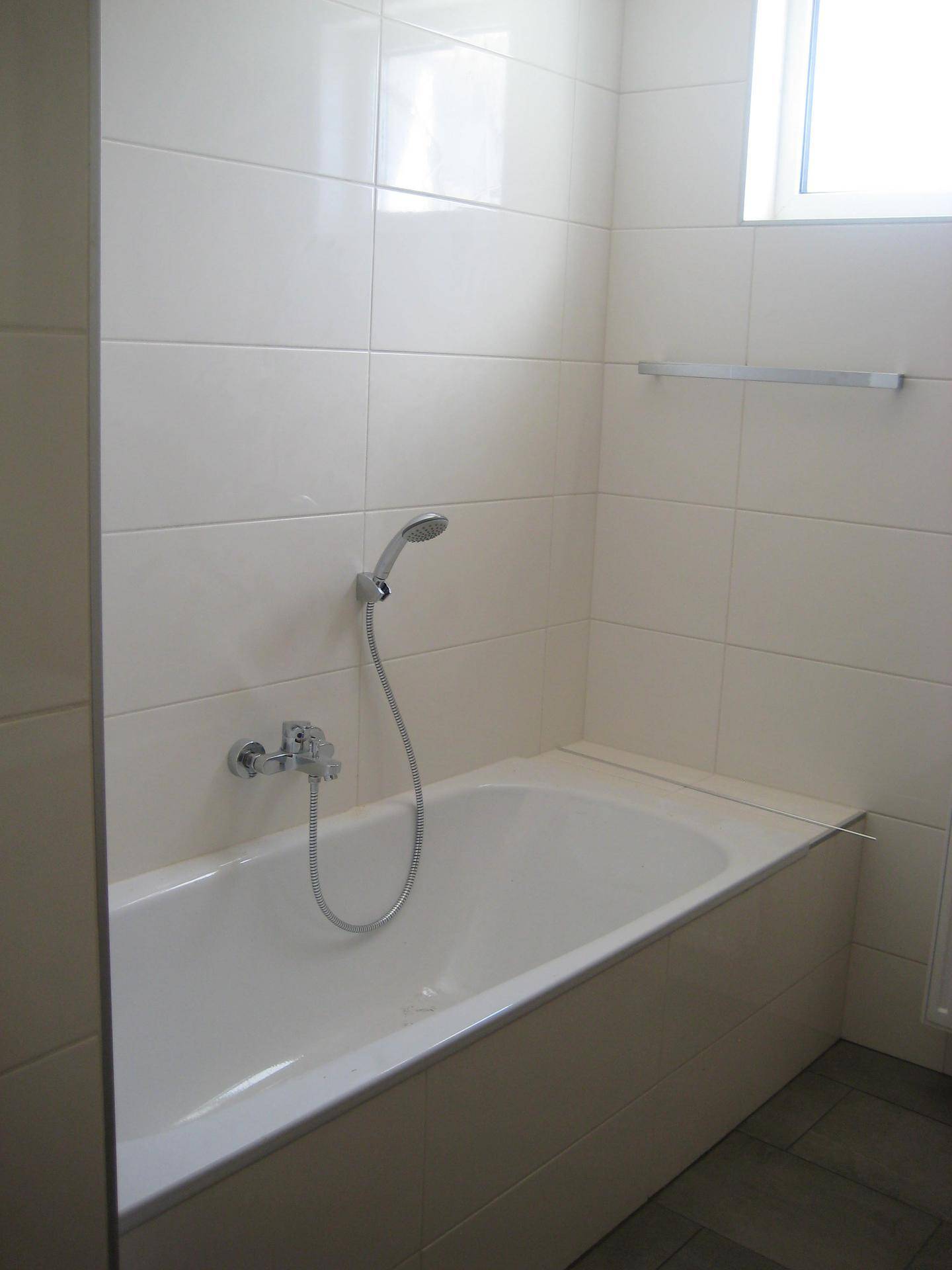 Gefliestes Badezimmer mit Badewanne und Duschvorrichtung, hell durch Fenster.