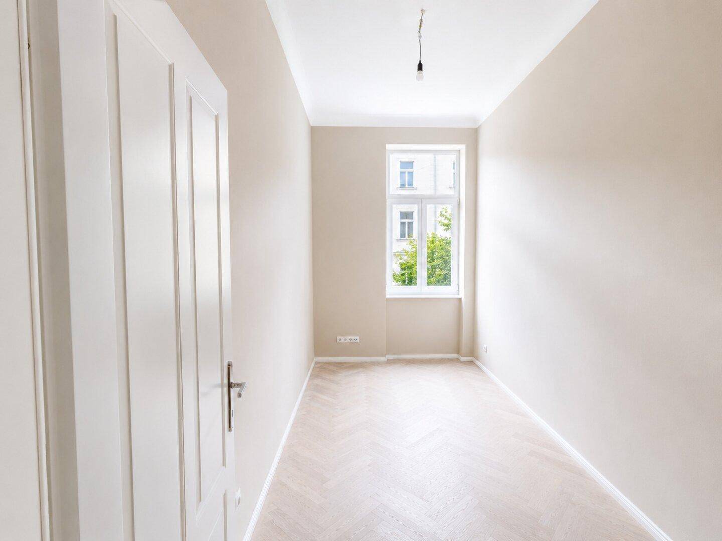 Helles Zimmer mit Fischgrätparkett und einem Fenster, das einen ruhigen Ausblick bietet.