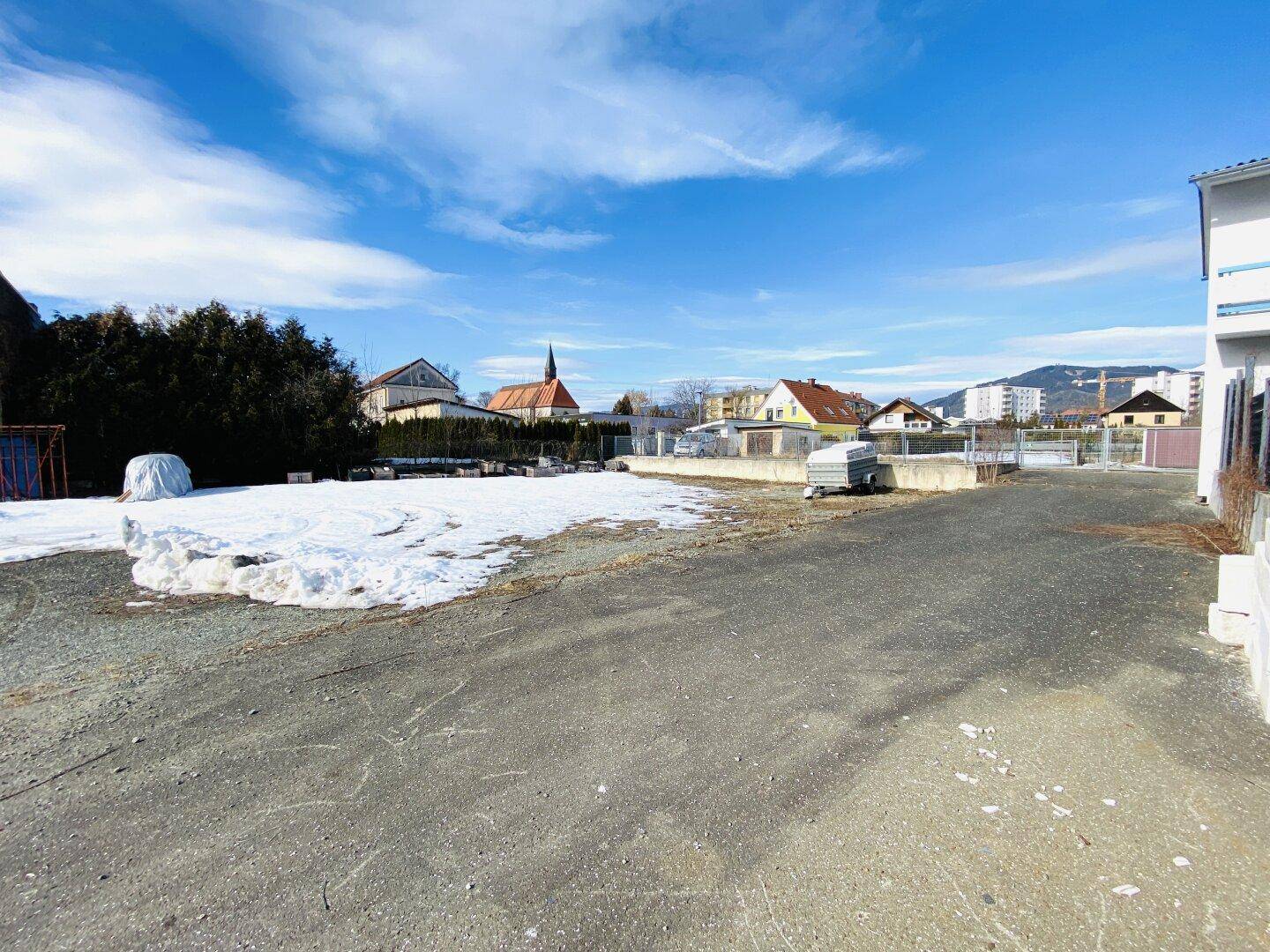 Großer, asphaltierter Außenbereich mit Schneeresten, umgeben von Wohngebäuden und Bergen im Hintergrund.