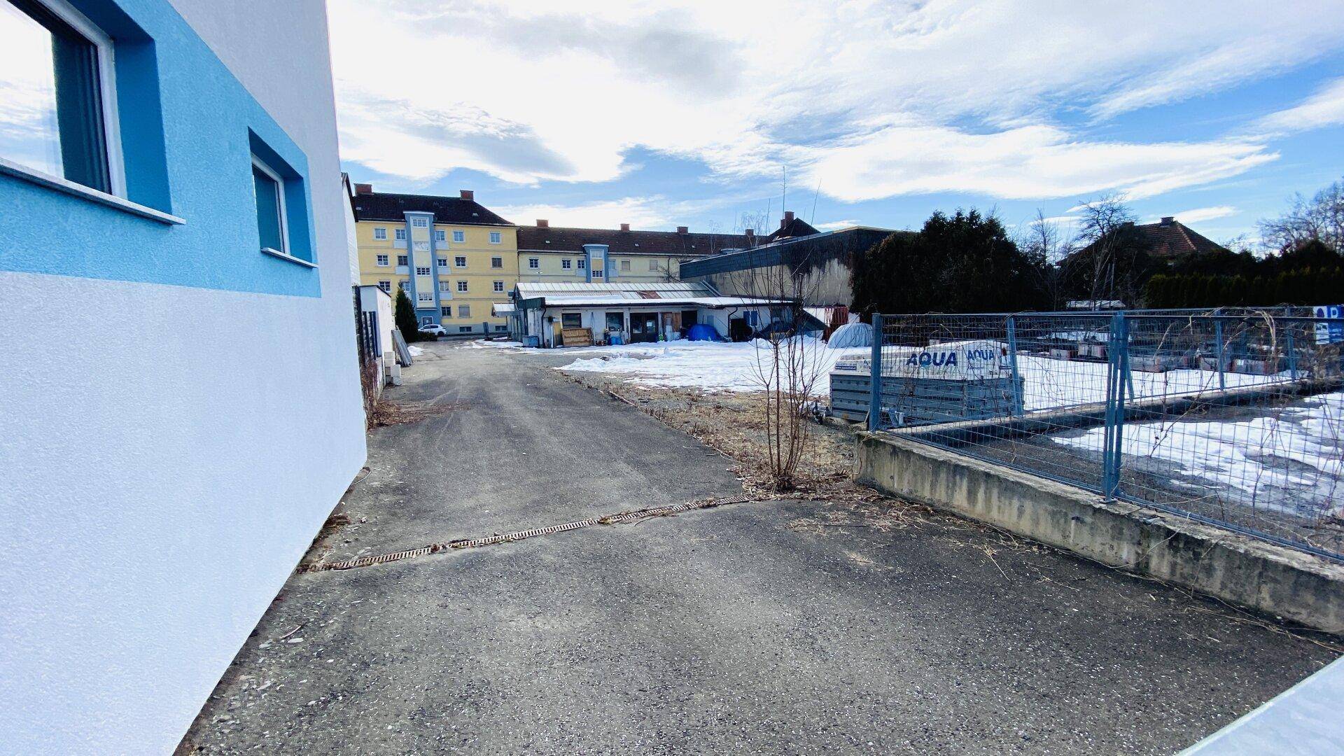 Gepflasterter Bereich neben einem Gebäude mit blau-weißer Fassade, Blick auf ein eingezäuntes Grundstück.