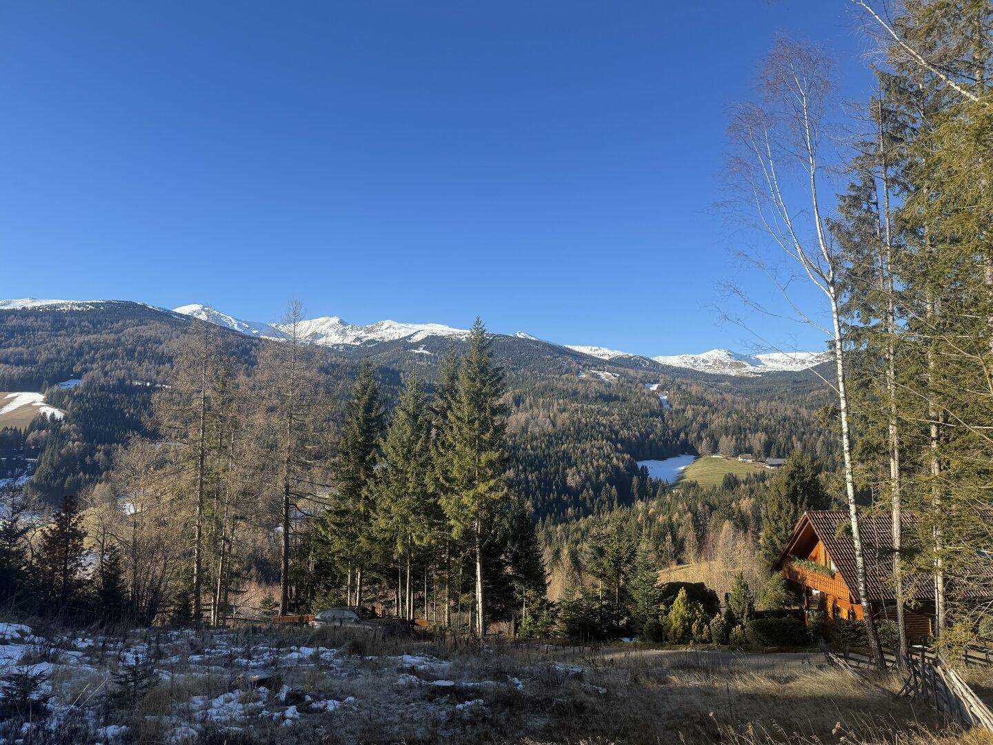 Weitläufige Landschaft mit bewaldeten Hügeln, schneebedeckten Gipfeln und einem kleinen Haus.