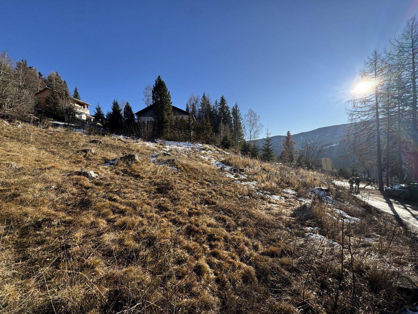 Sonnige Hanglandschaft mit trockenem Gras, Bäumen und Häusern am Horizont.