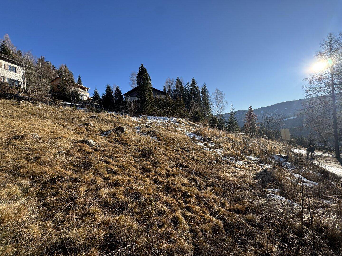 Weitläufiges Hanggrundstück mit Gras, Schnee und Bäumen, umgeben von ländlicher Bebauung.