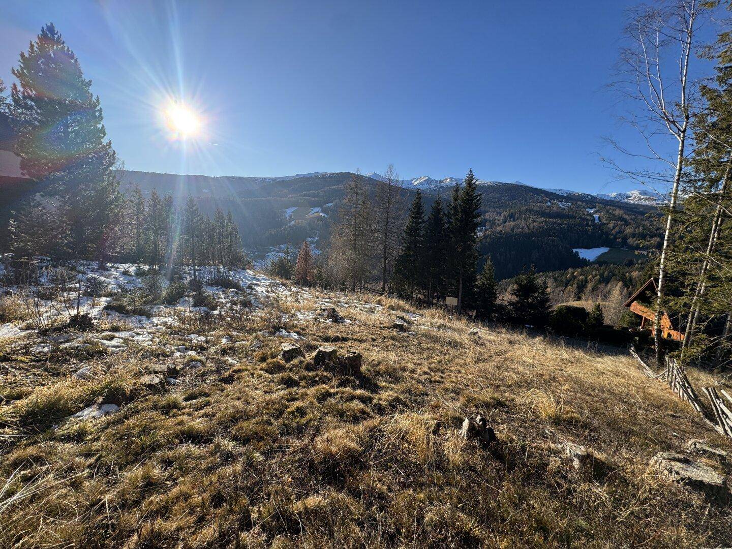 Sonnige Landschaft mit weiten Hügeln, Wäldern und schneebedeckten Berggipfeln im Hintergrund.