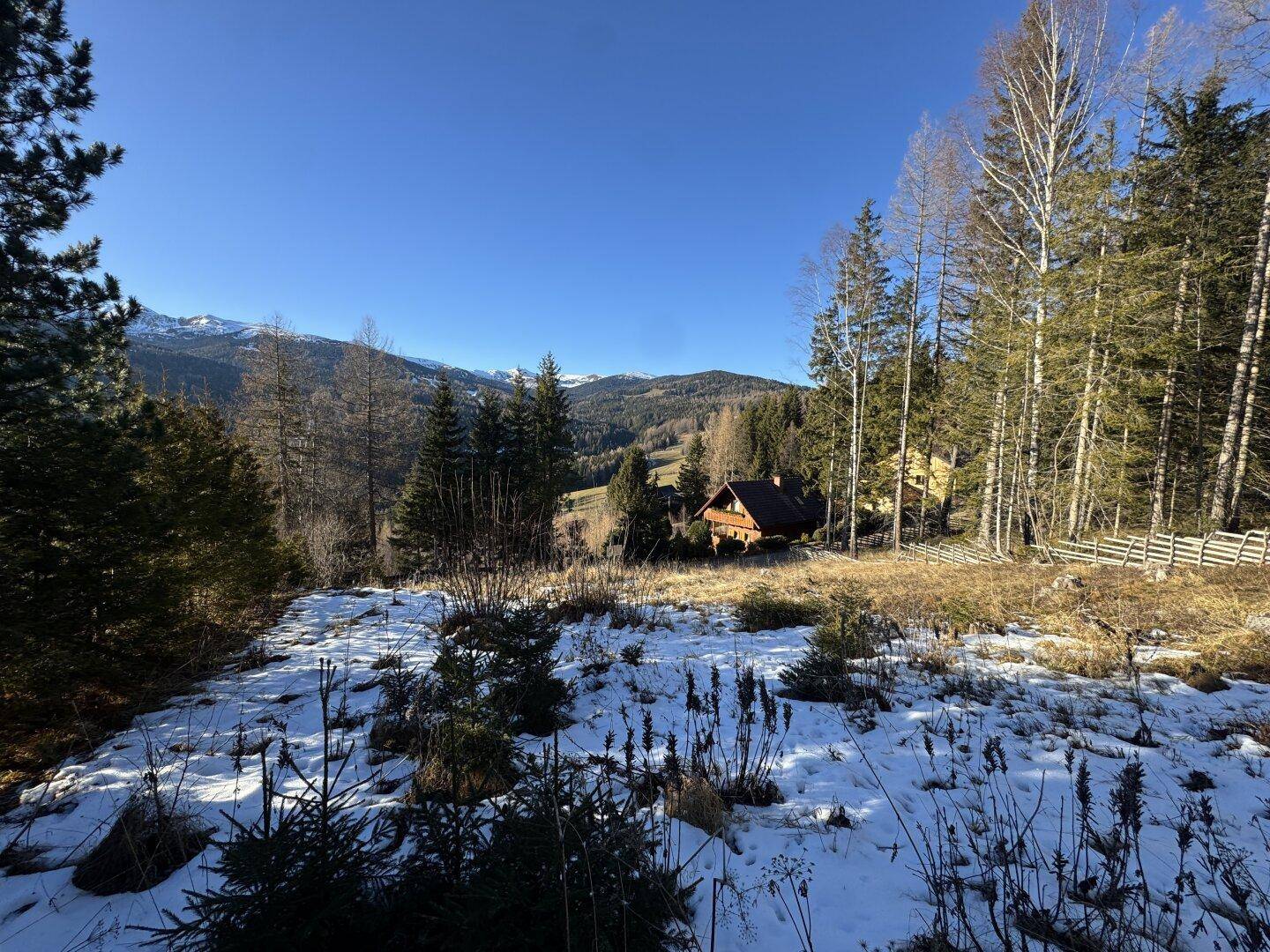 Idyllisches Holzhaus umgeben von Bäumen und Schnee mit majestätischen Bergen im Hintergrund.