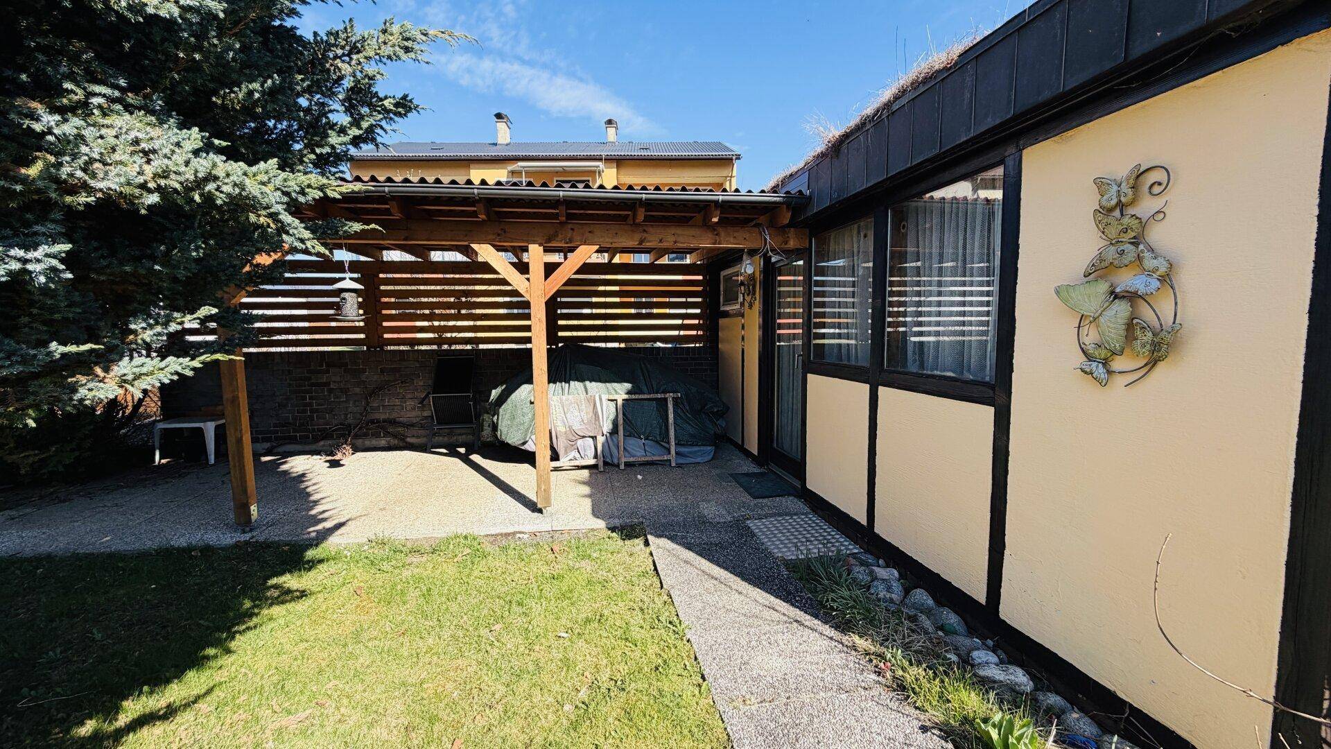 Überdachte Terrasse mit Holzbalken und Zugang zu einer kleinen Rasenfläche am Haus.
