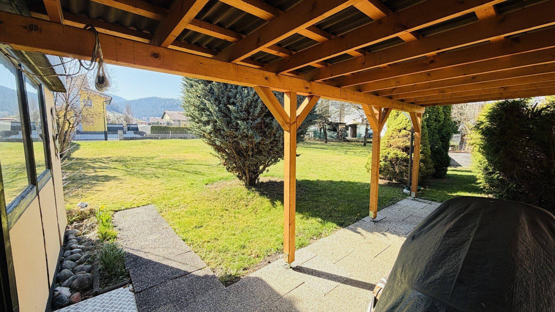Blick von der überdachten Terrasse auf die weitläufige grüne Gartenfläche mit Bäumen.