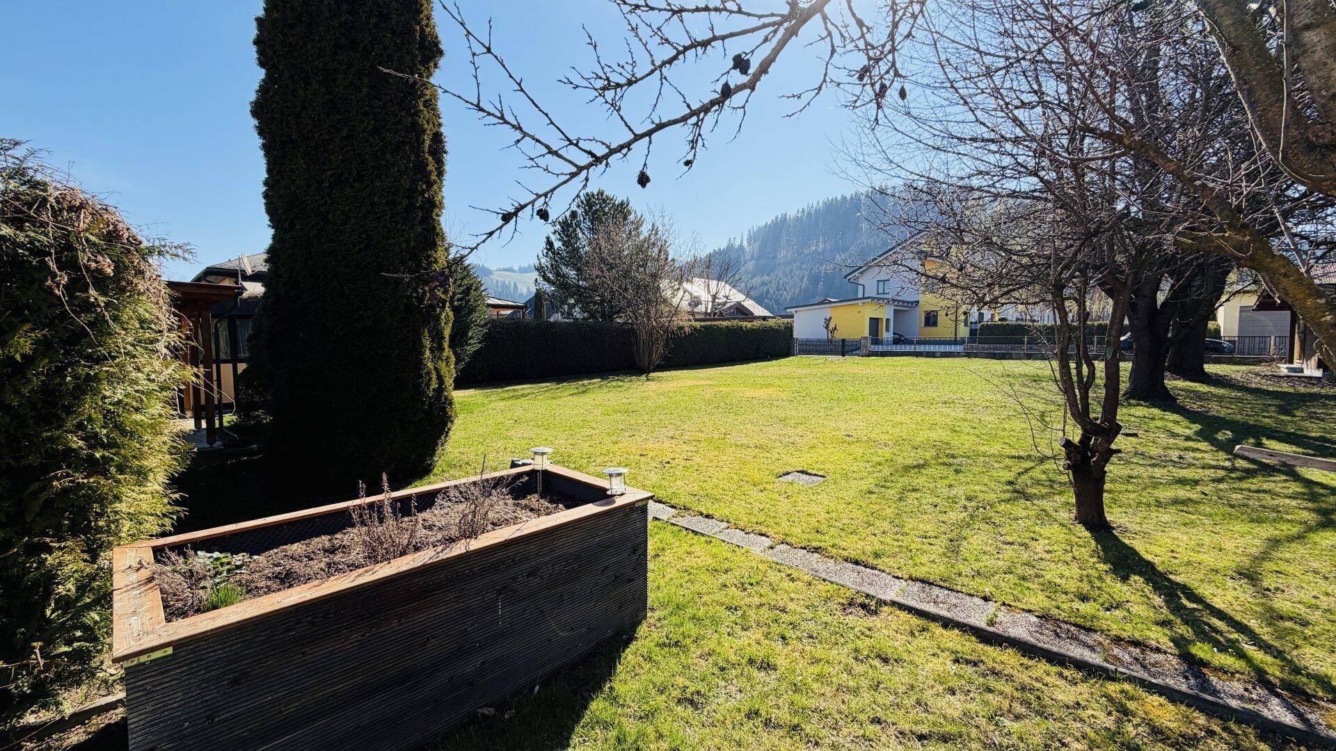 Gartenansicht mit einem erhöhten Pflanzbeet, Bäumen und Häusern in der Ferne.