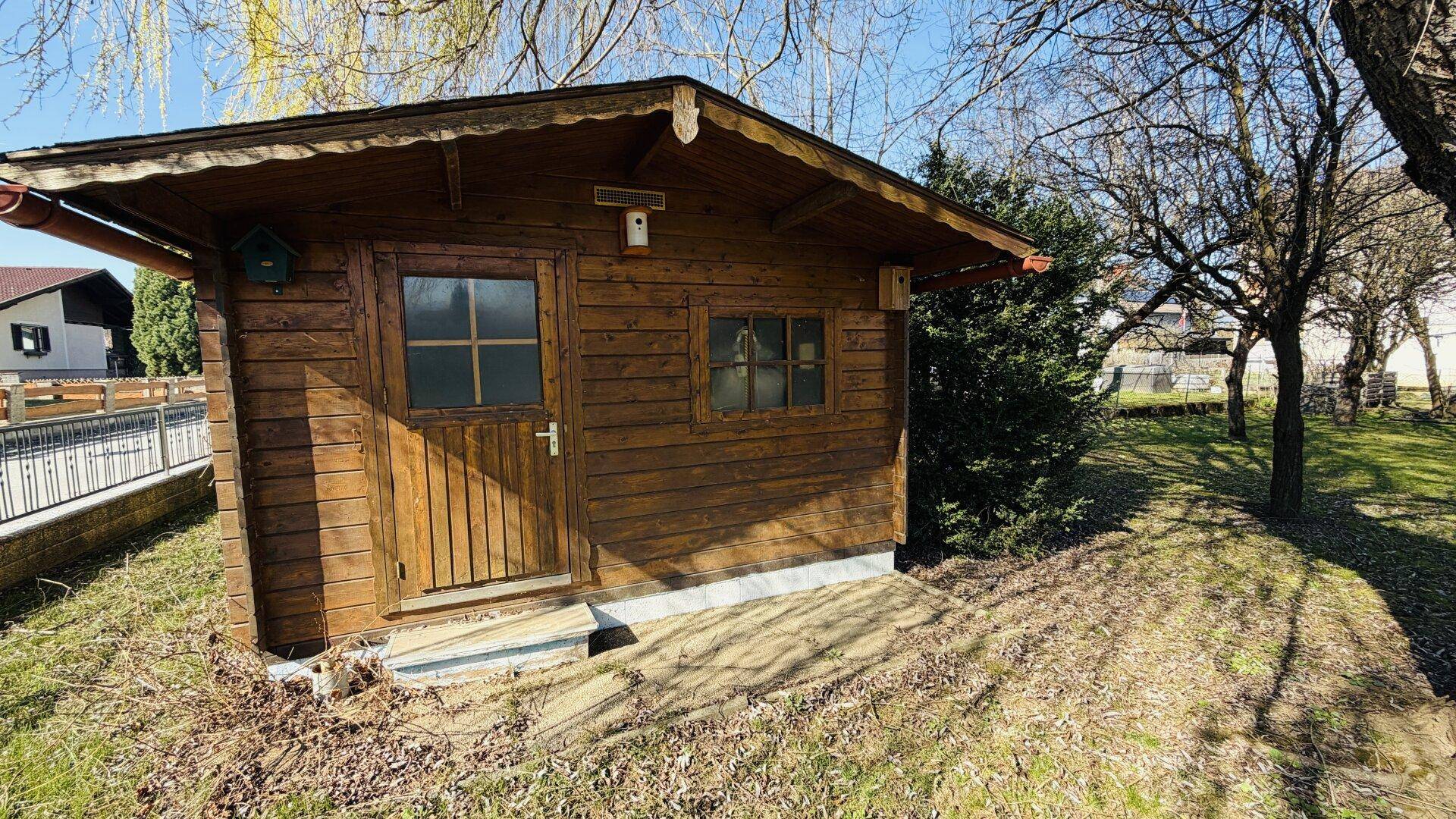 Rustikaler Holzschuppen mit Fenstern und einer Tür, umgeben von Gras und Bäumen.