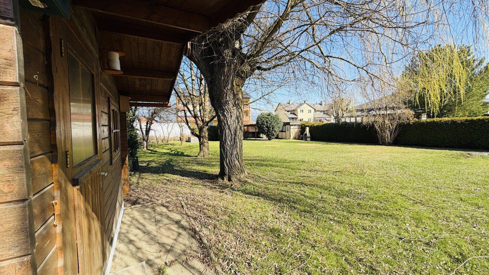 Blick vom Holzschuppen über die weite Rasenfläche mit Bäumen und Hecken.