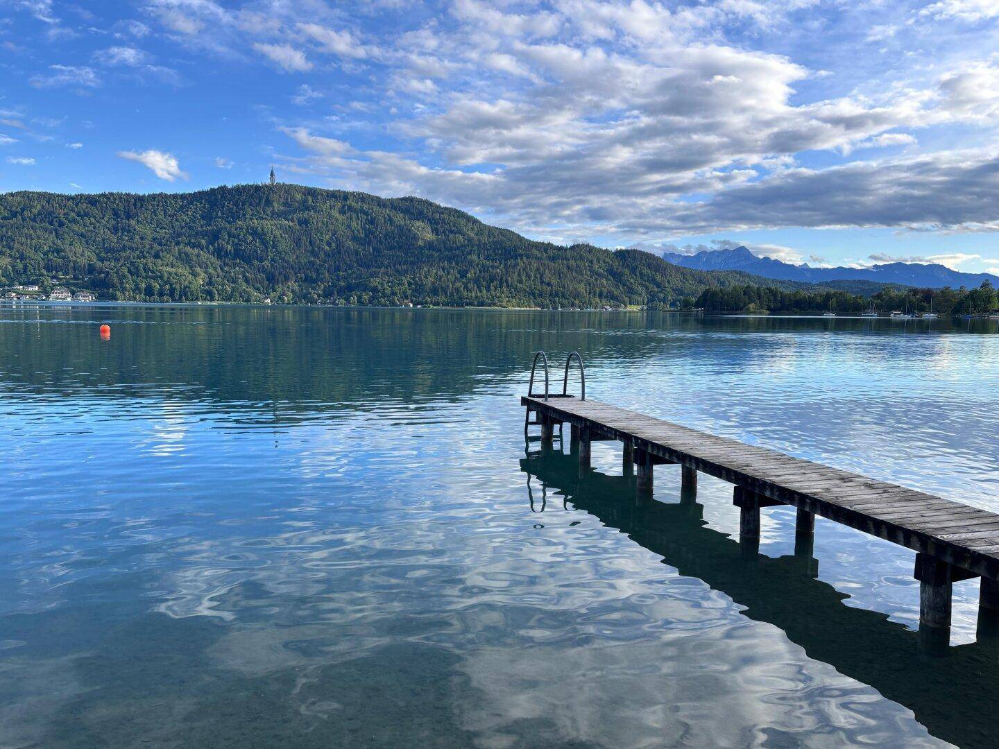 Malerischer See mit Holzsteg und bewaldeten Bergen im Hintergrund unter blauem Himmel.