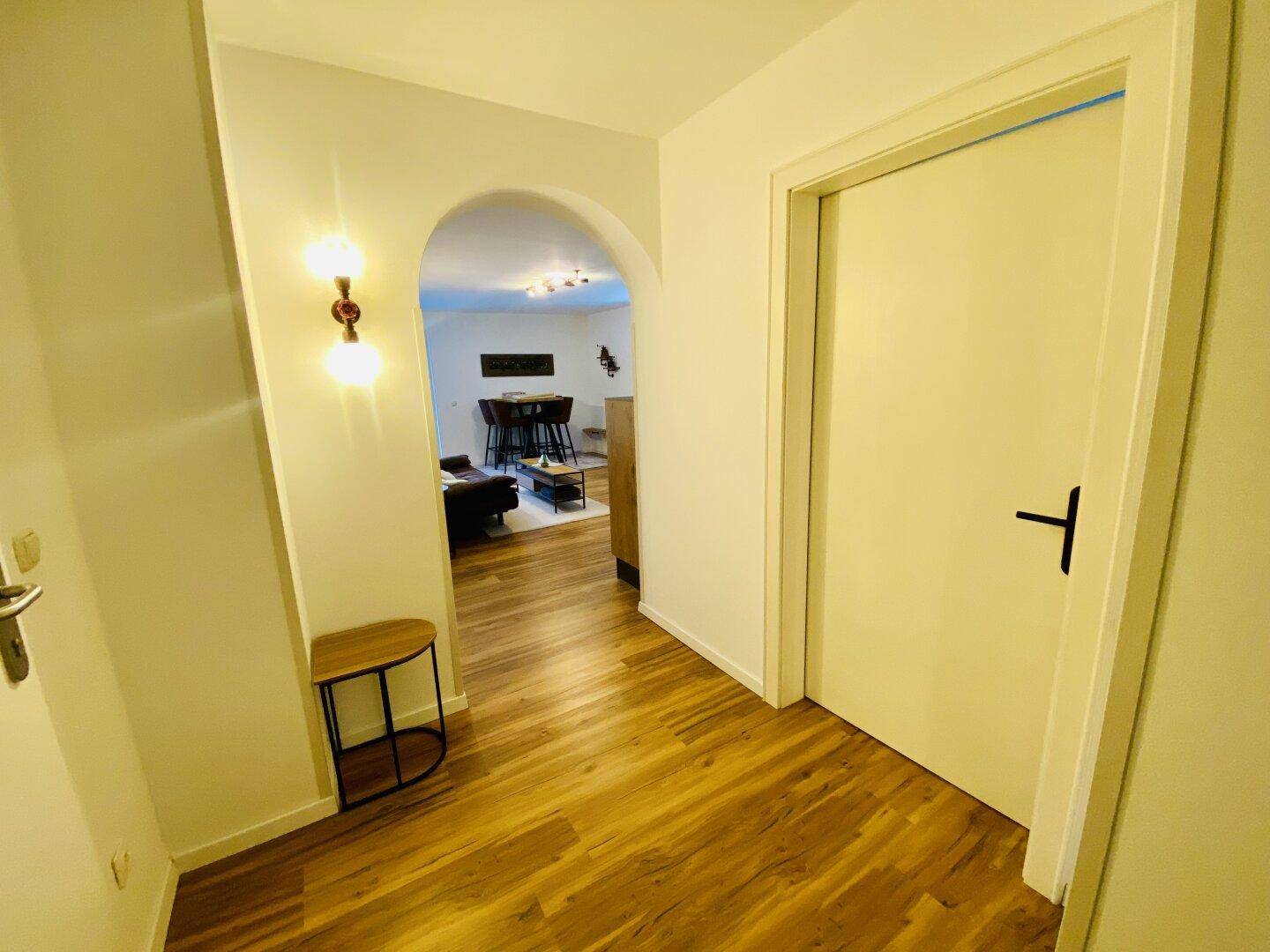 Eingangsbereich mit Holzboden, Wandleuchte und Blick durch einen Bogen in den Wohnbereich.