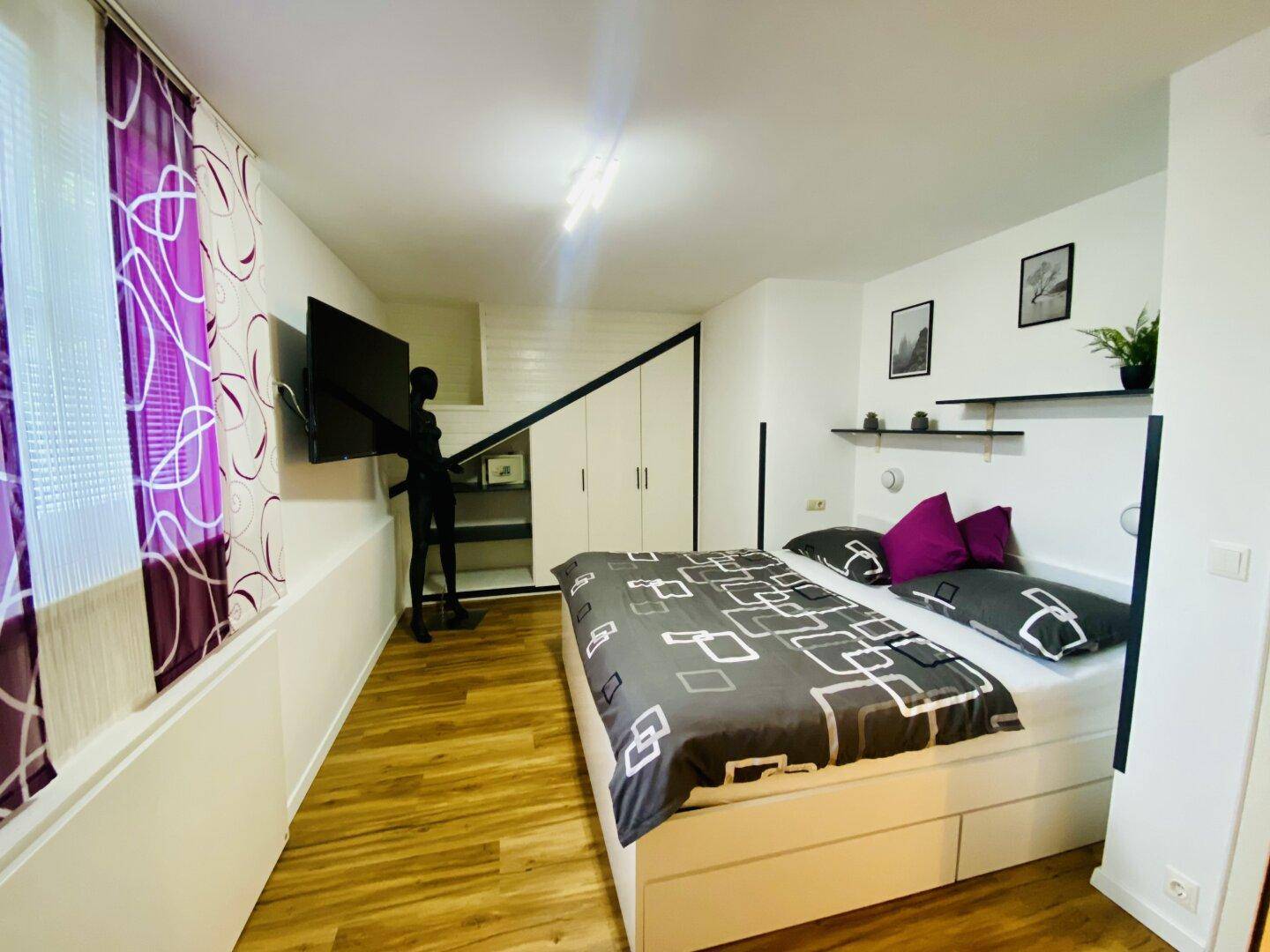 Helles Schlafzimmer mit Holzboden, Doppelbett, Wandfernseher und modernen Regalen.