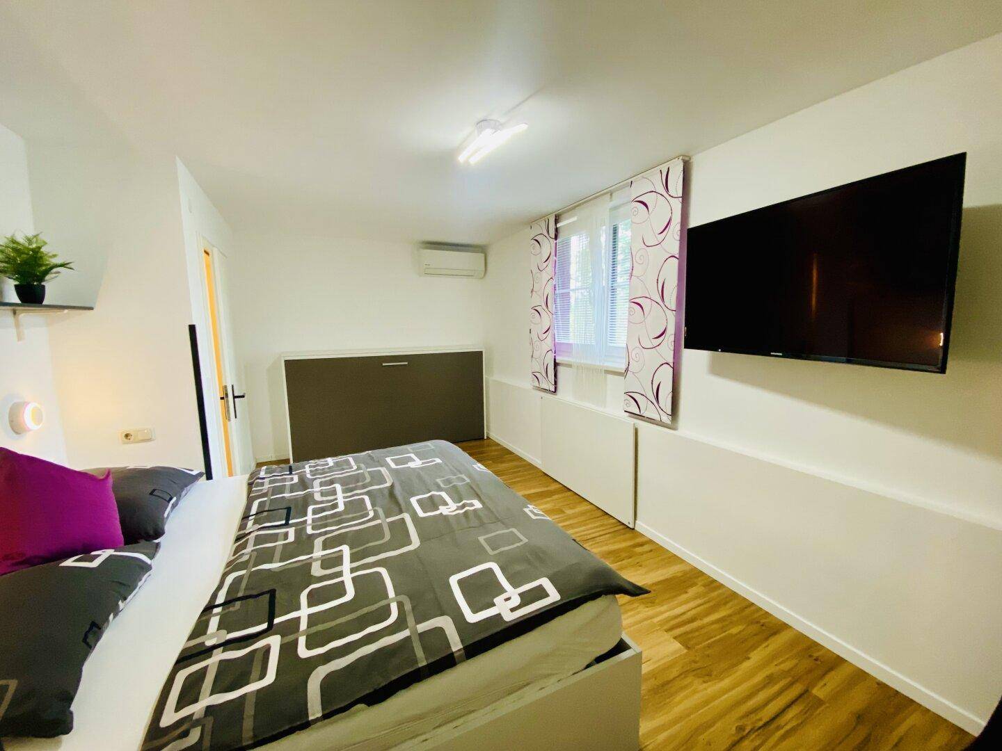 Helles Schlafzimmer mit Holzboden, Doppelbett, Wandfernseher und Klimaanlage.