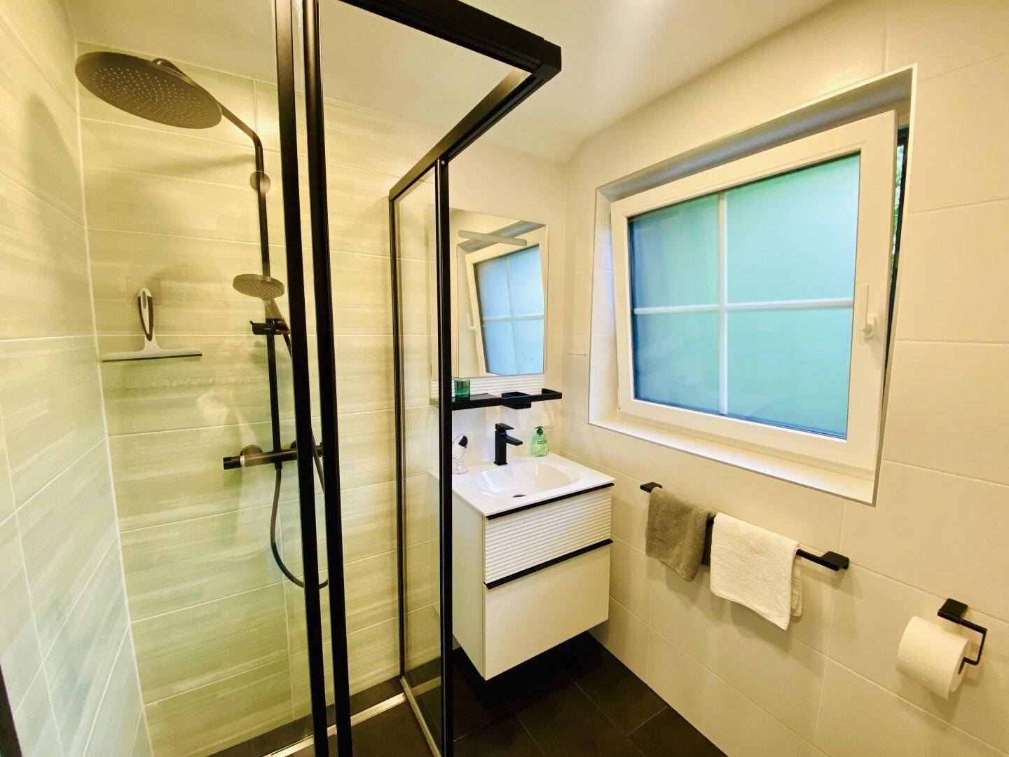 Helles Badezimmer mit moderner Duschkabine, Waschtisch und Fenster.