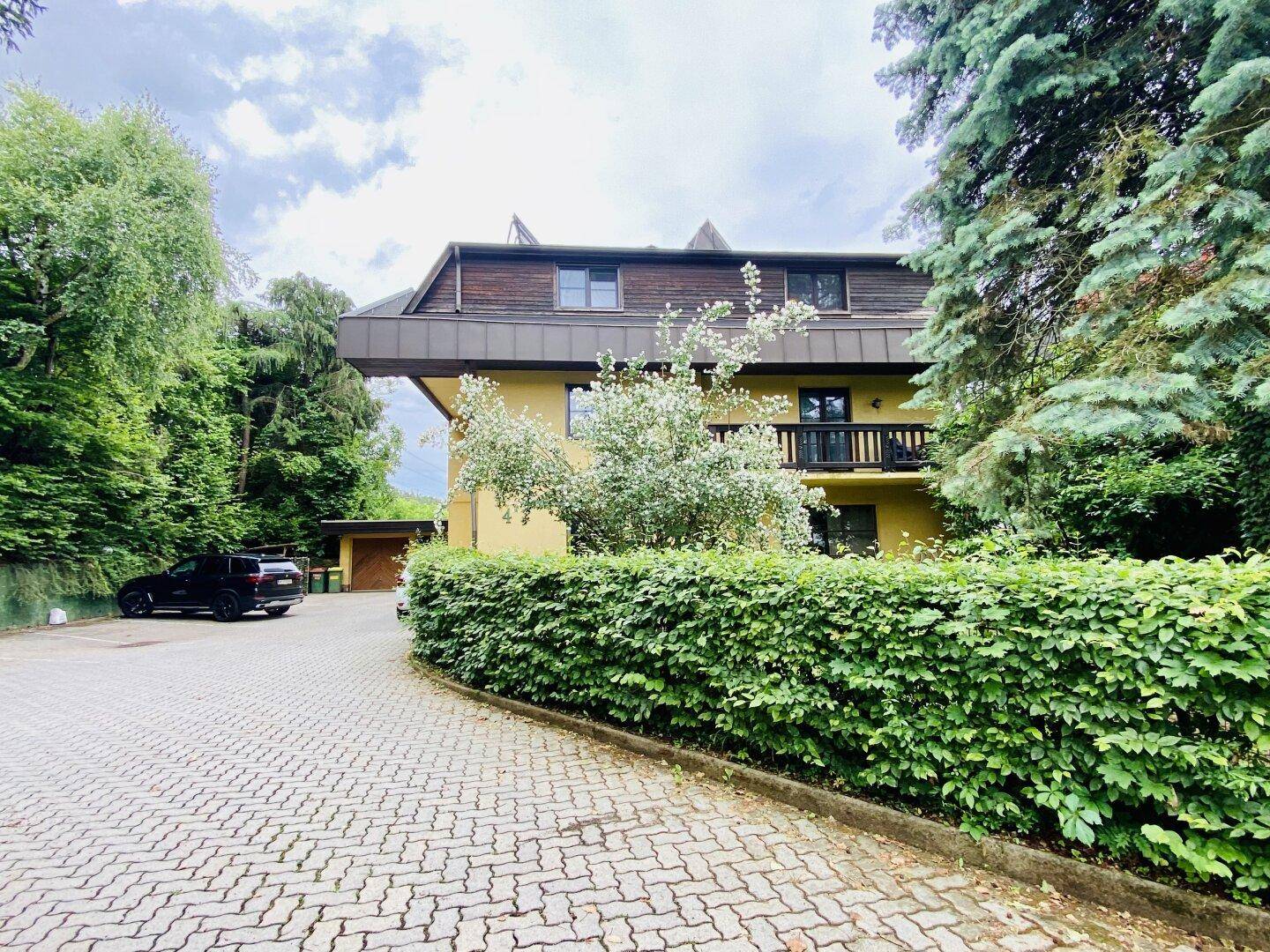 Mehrfamilienhaus mit gelber Fassade, gepflasterter Auffahrt und grüner Hecke.