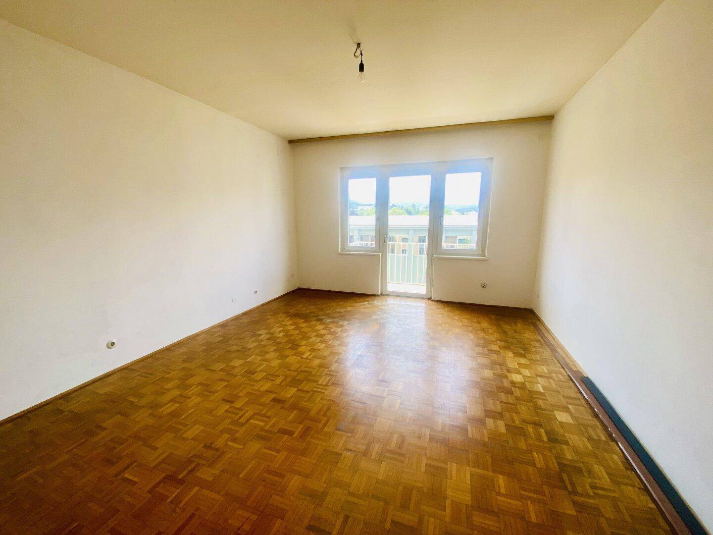 Helles Wohnzimmer mit Fischgrätparkettboden und Zugang zu einem kleinen Balkon.