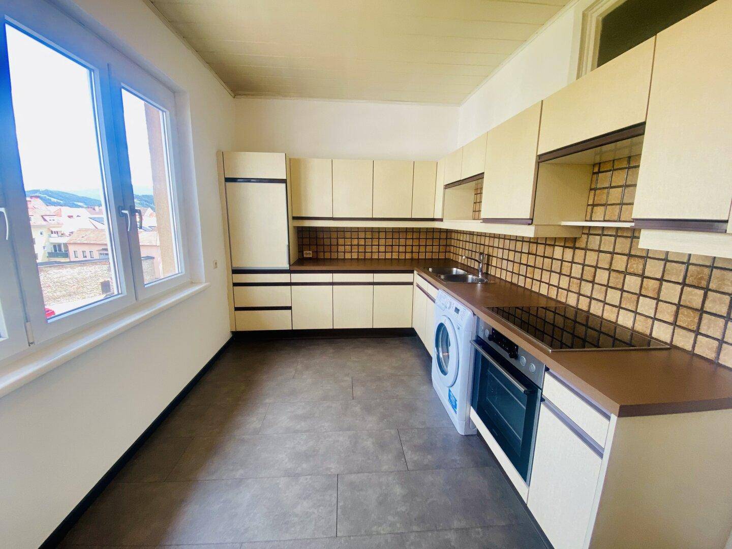 Funktionale Küche mit älteren Einbauschränken, Fliesenboden und großem Fenster für viel Licht.