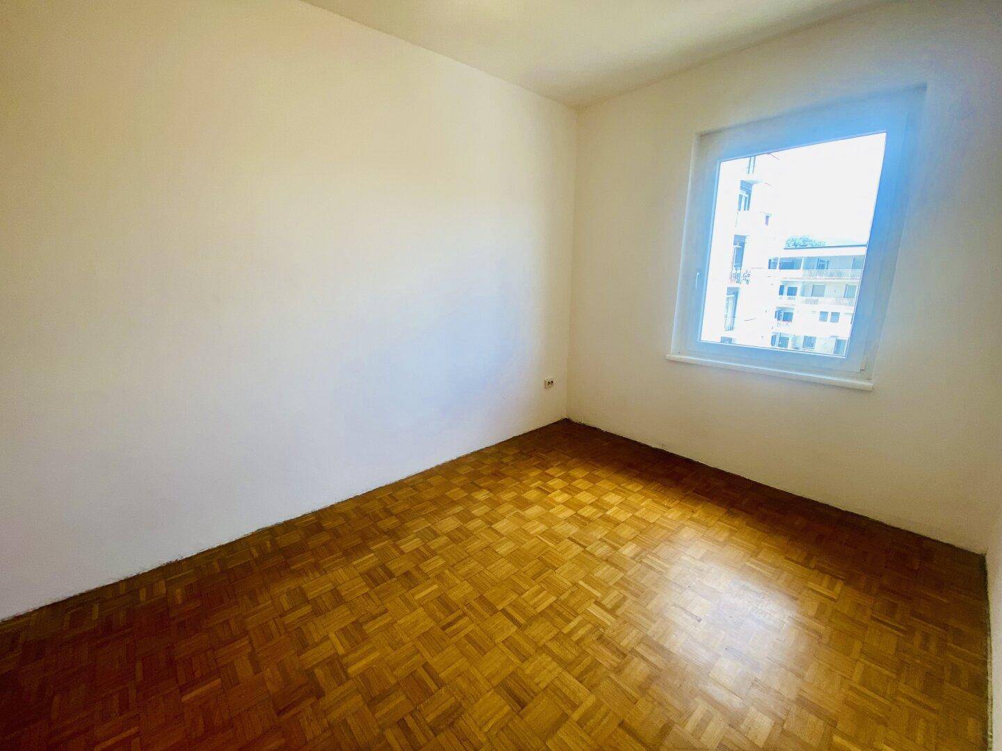 Helles Schlafzimmer mit Fischgrätparkettboden und einem Fenster, das viel Tageslicht bietet.