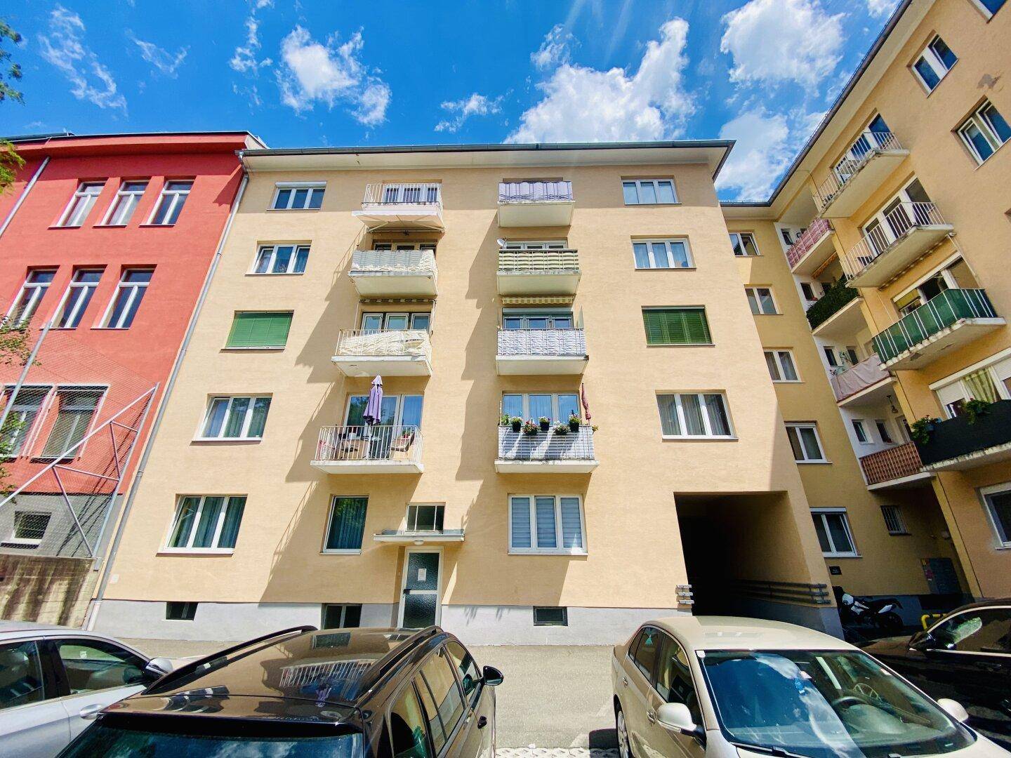 Fassade eines Mehrfamilienhauses mit mehreren Balkonen und Parkplätzen davor.