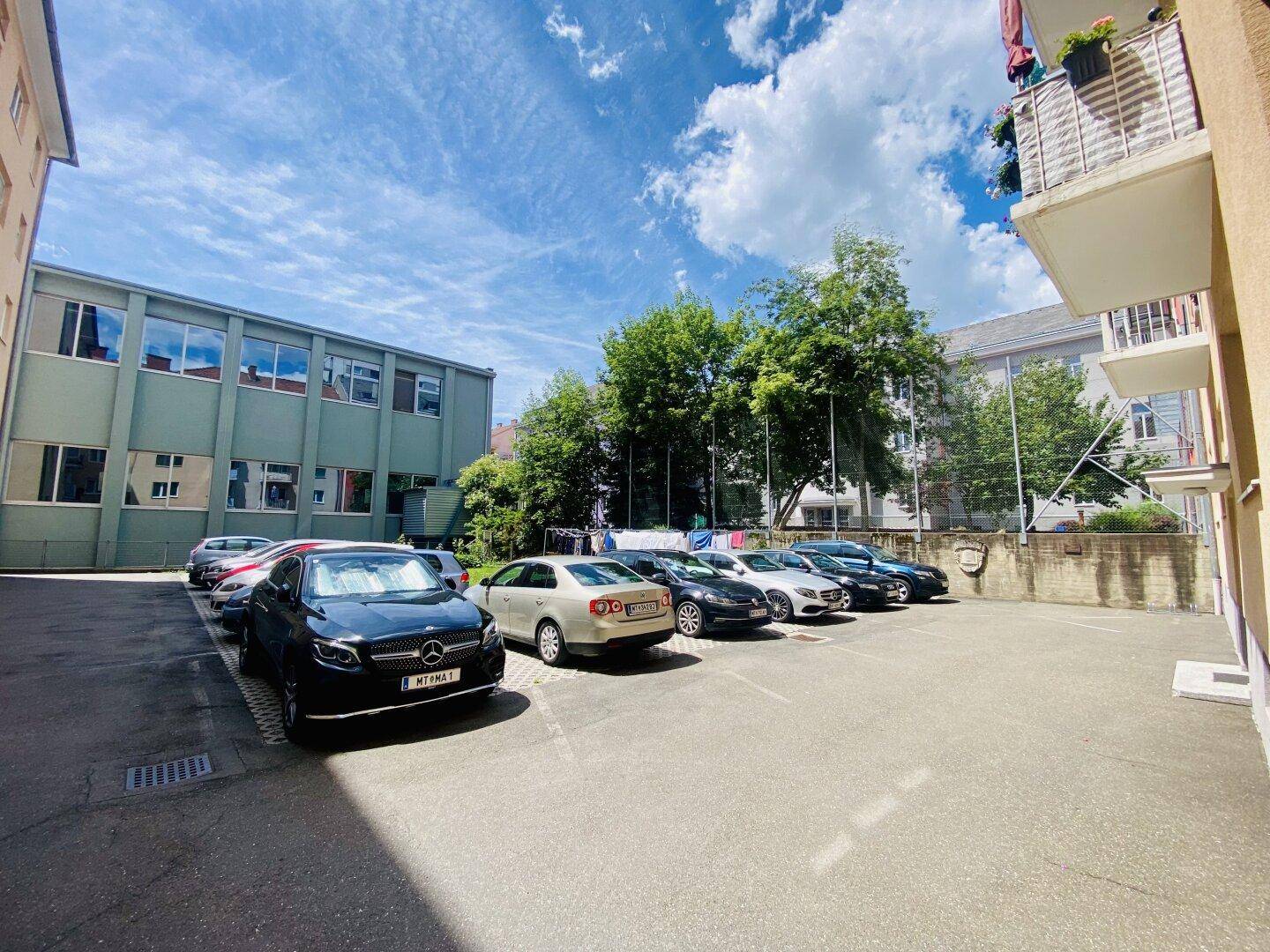 Außenparkplatz mit mehreren geparkten Fahrzeugen und umliegenden Gebäuden unter blauem Himmel.