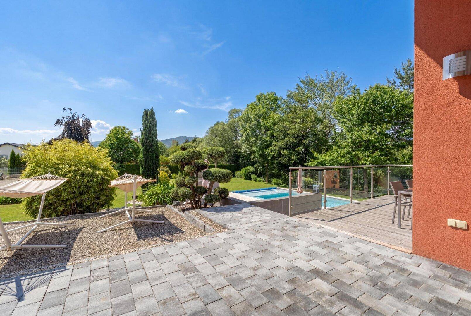 Große Terrasse mit Steinplatten und Holzdeck, umgeben von Grünflächen und Blick auf den Pool.