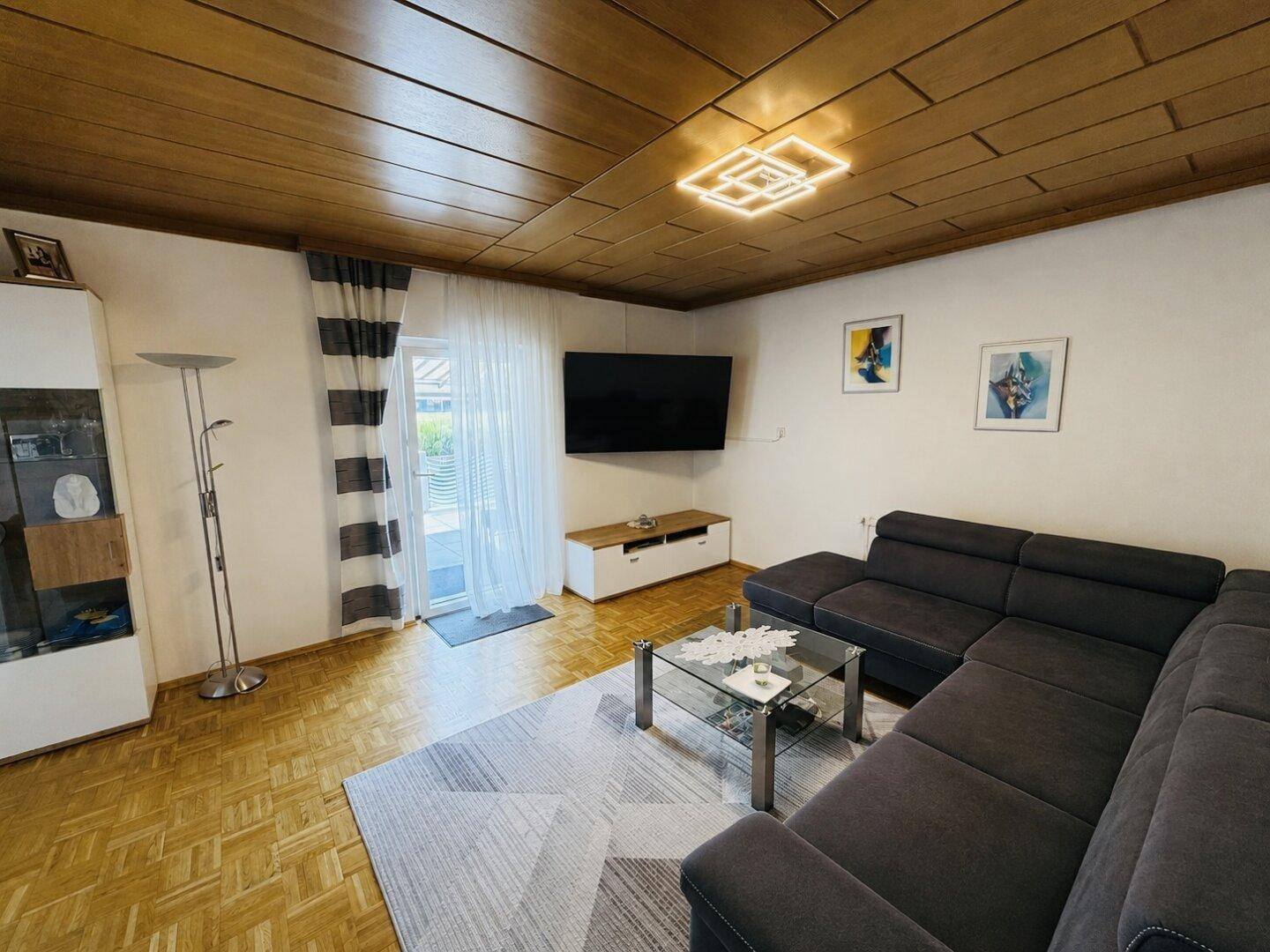 Wohnzimmer mit Holzkassettendecke, Ecksofa und Zugang zum Außenbereich.