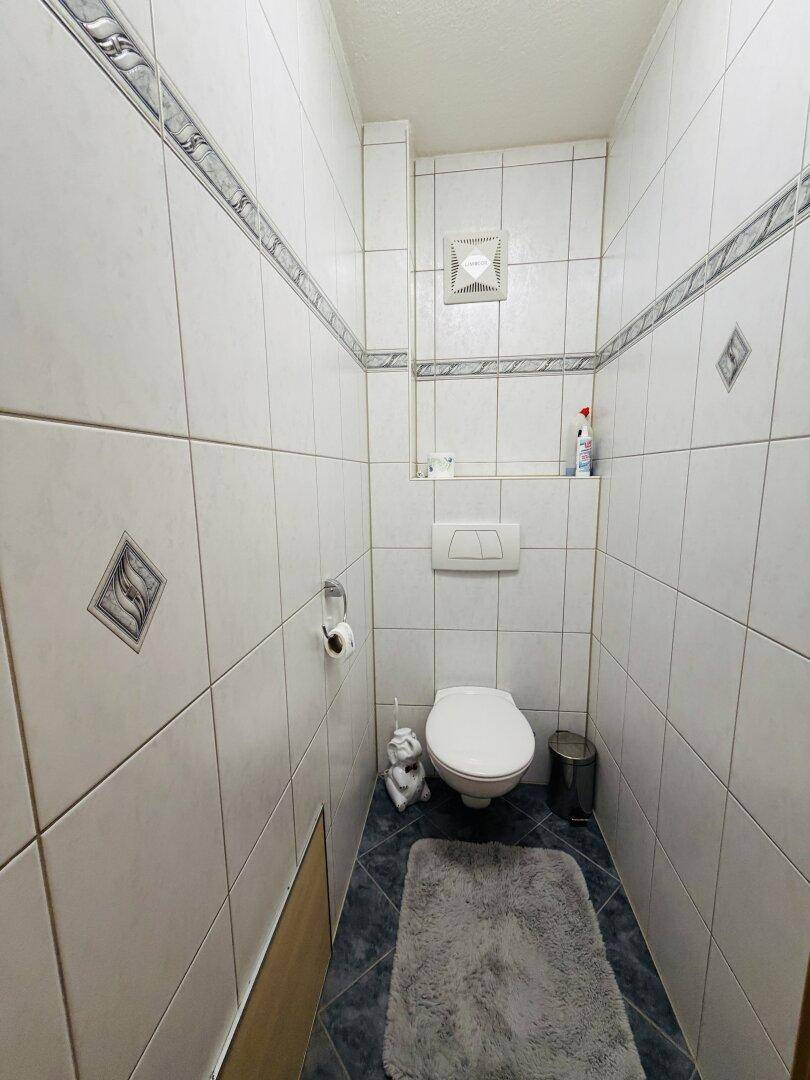 Kleines Gäste-WC mit Wand-WC, Fliesenboden und Abluftventilator an der Decke.