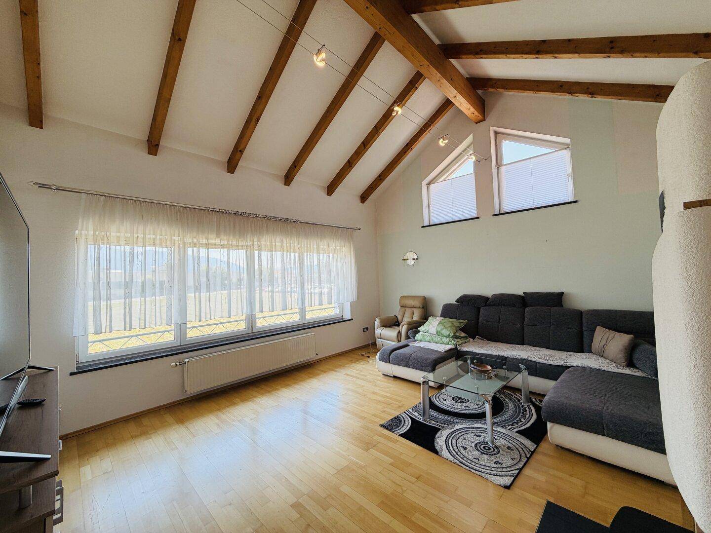 Gemütliches Wohnzimmer mit Holzbalkendecke, bequemer Couch und viel Tageslicht durch große Fenster.