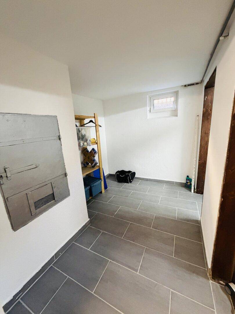 Kellerraum mit gefliestem Boden, kleinem Fenster und einfacher Einrichtung für Lagerzwecke.