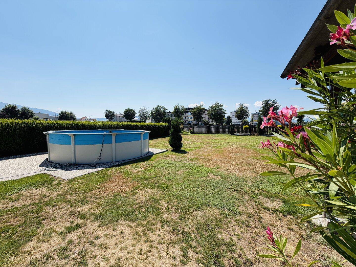 Sonniger Gartenbereich mit Aufstellpool, gepflegter Hecke und blühenden Pflanzen im Vordergrund.
