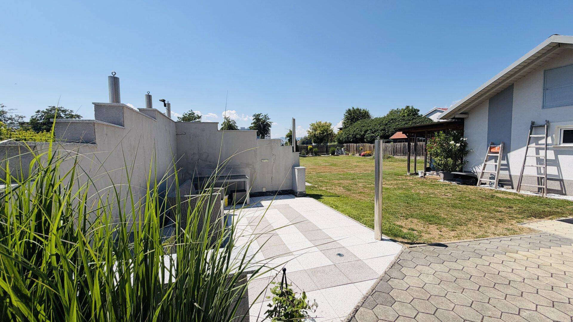 Gepflasterter Bereich neben dem Haus, umgeben von Grünflächen und einer Pergola im Hintergrund.