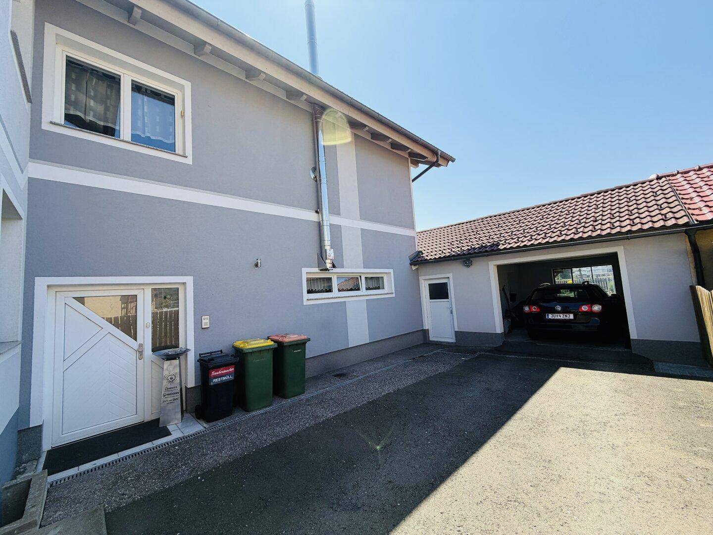 Seitenansicht des Hauses mit Eingangsbereich, Mülltonnen und offener Garage mit Fahrzeug.
