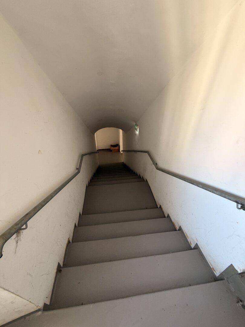 Langer, schmaler Treppenabgang mit Metallhandläufen, der in einen dunkleren Bereich führt.