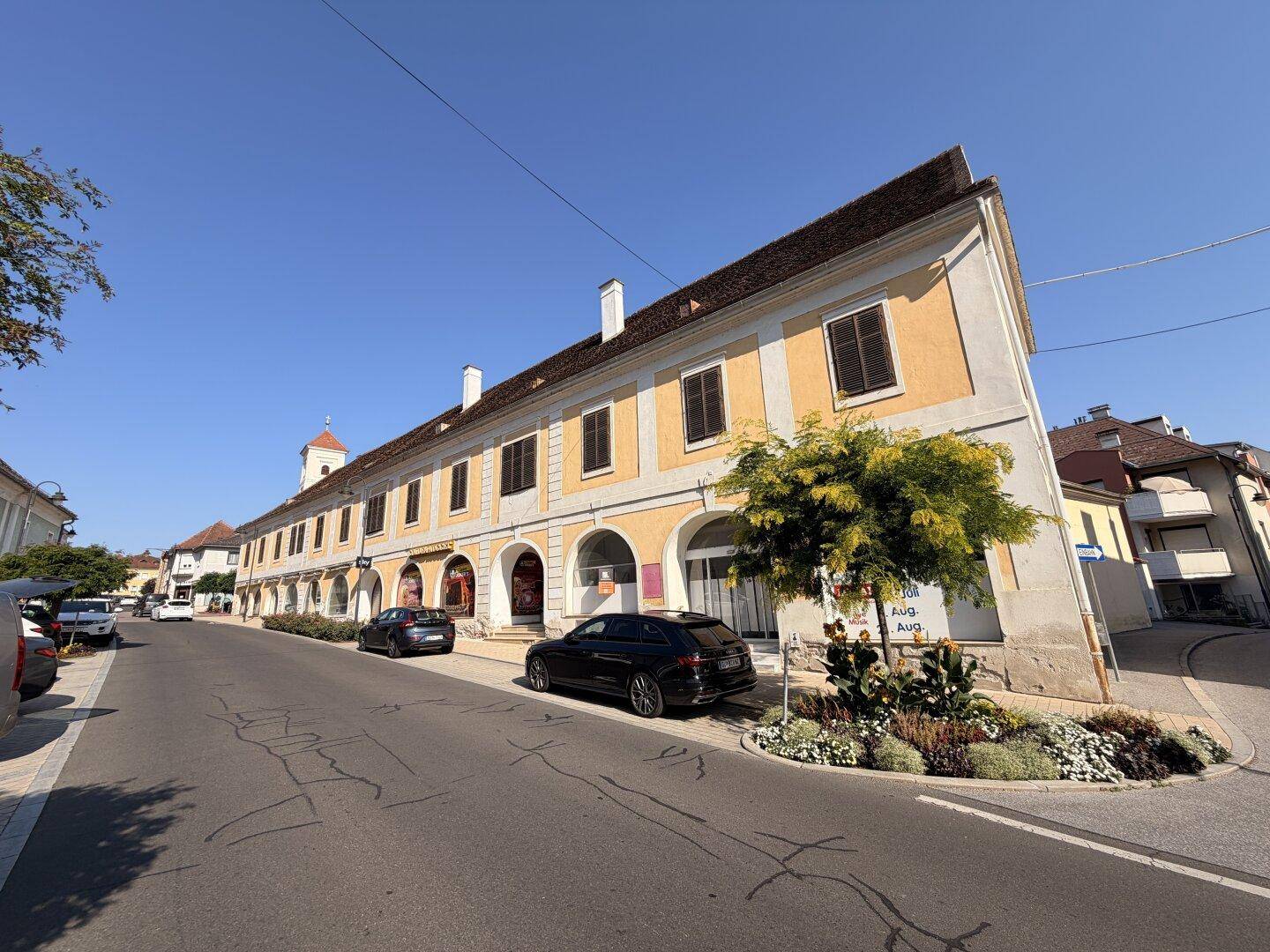 Sonnige Straßenansicht eines langen, historischen Gebäudes mit Arkaden und geparkten Autos.