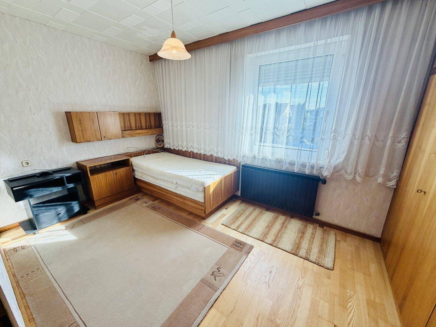 Schlafzimmer mit Einzelbett, Holzmöbeln und Parkettboden, hell durch großes Fenster.