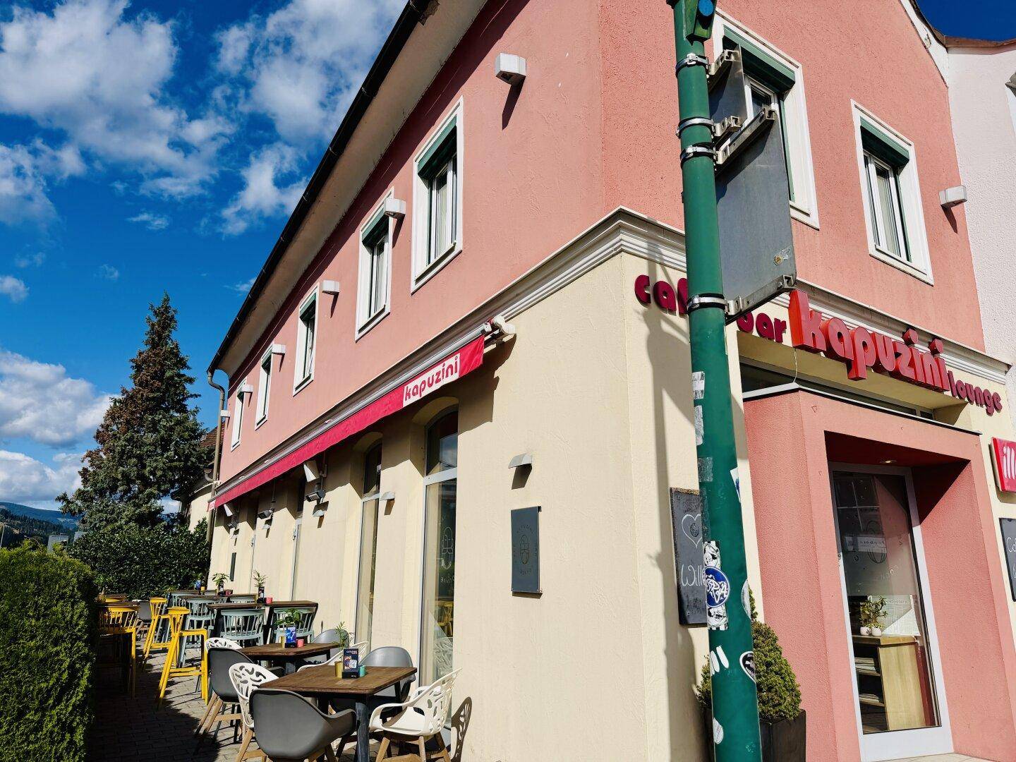 Eckansicht eines Gebäudes mit rosafarbener Fassade und einer Außenterrasse mit Markise.