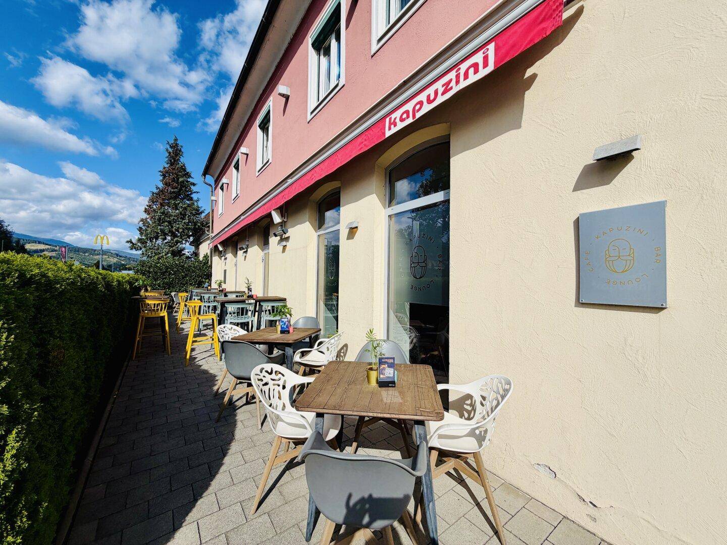 Eine gepflasterte Außenterrasse mit Tischen und Stühlen, umgeben von einer grünen Hecke und sonnigem Wetter.