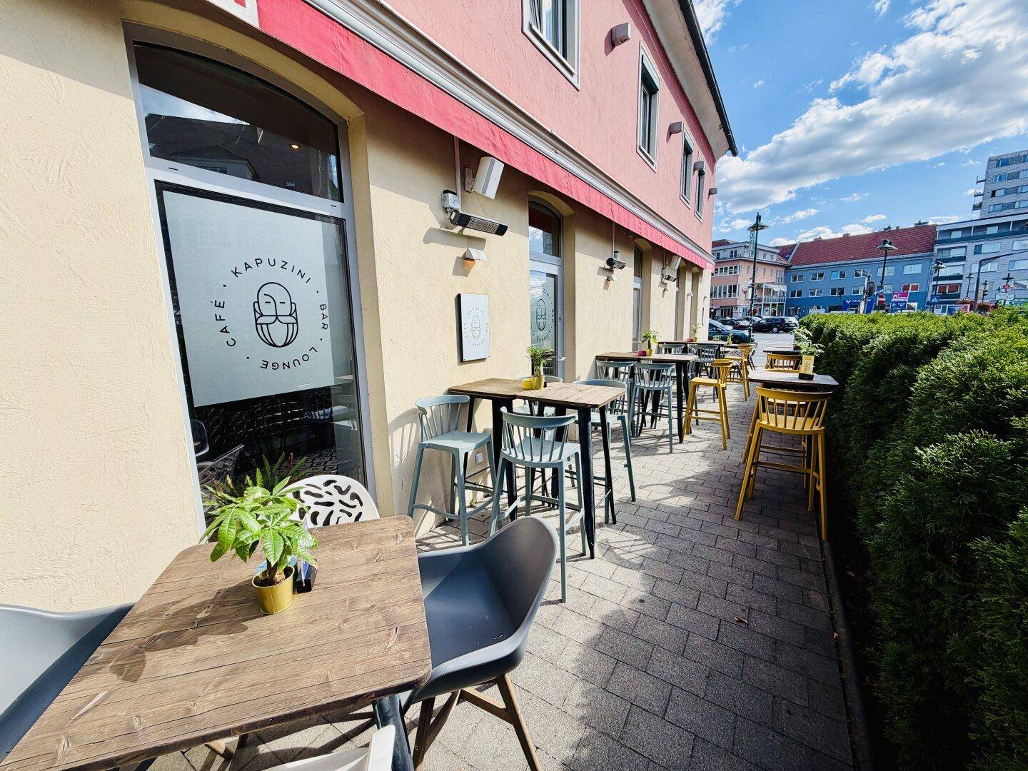 Eine sonnige Außenterrasse mit modernen Tischen und Stühlen vor der Gebäudefassade und einer Hecke.
