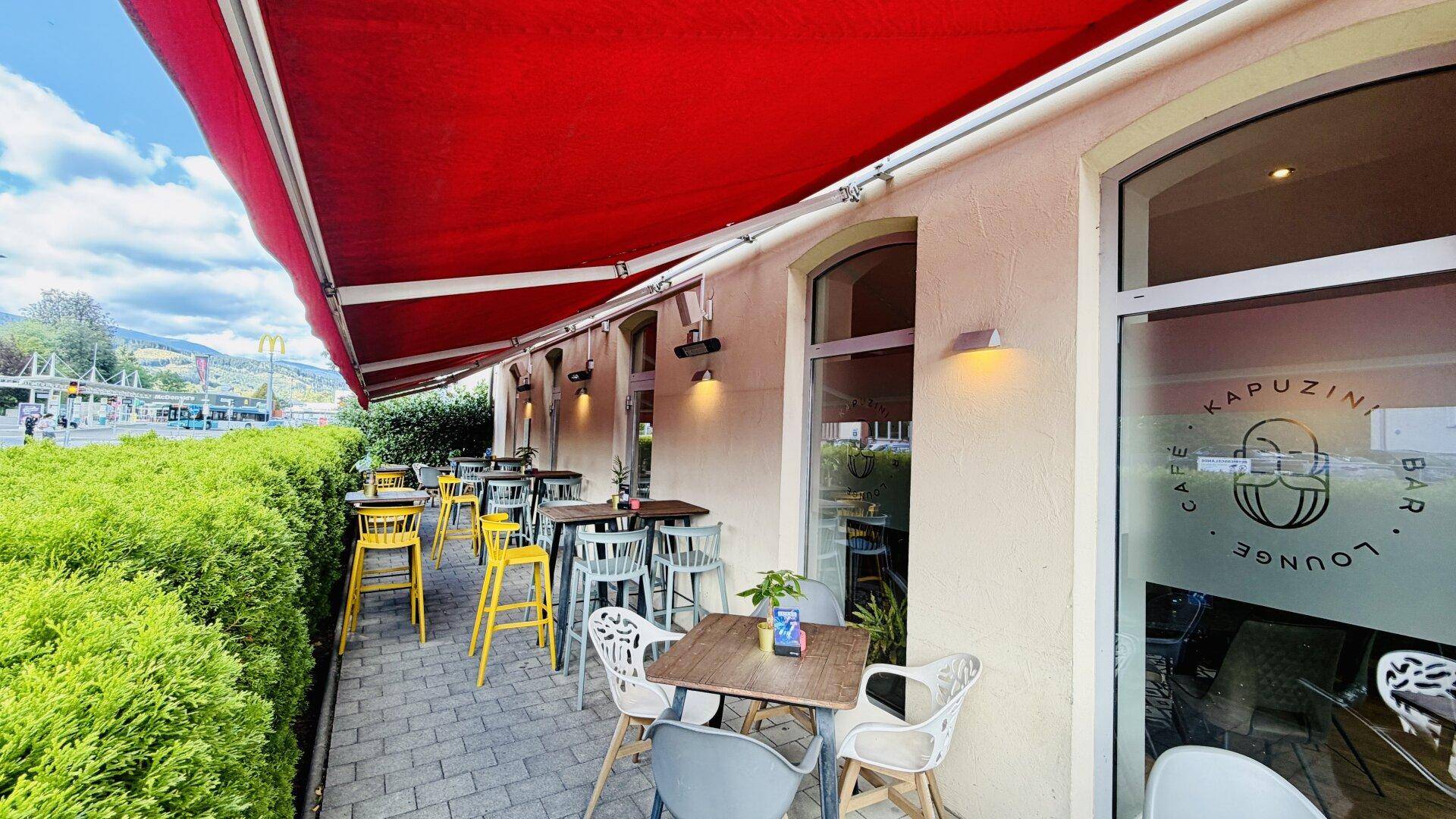 Eine sonnige Außenterrasse mit modernen Tischen und Stühlen, geschützt durch eine rote Markise und Hecke.