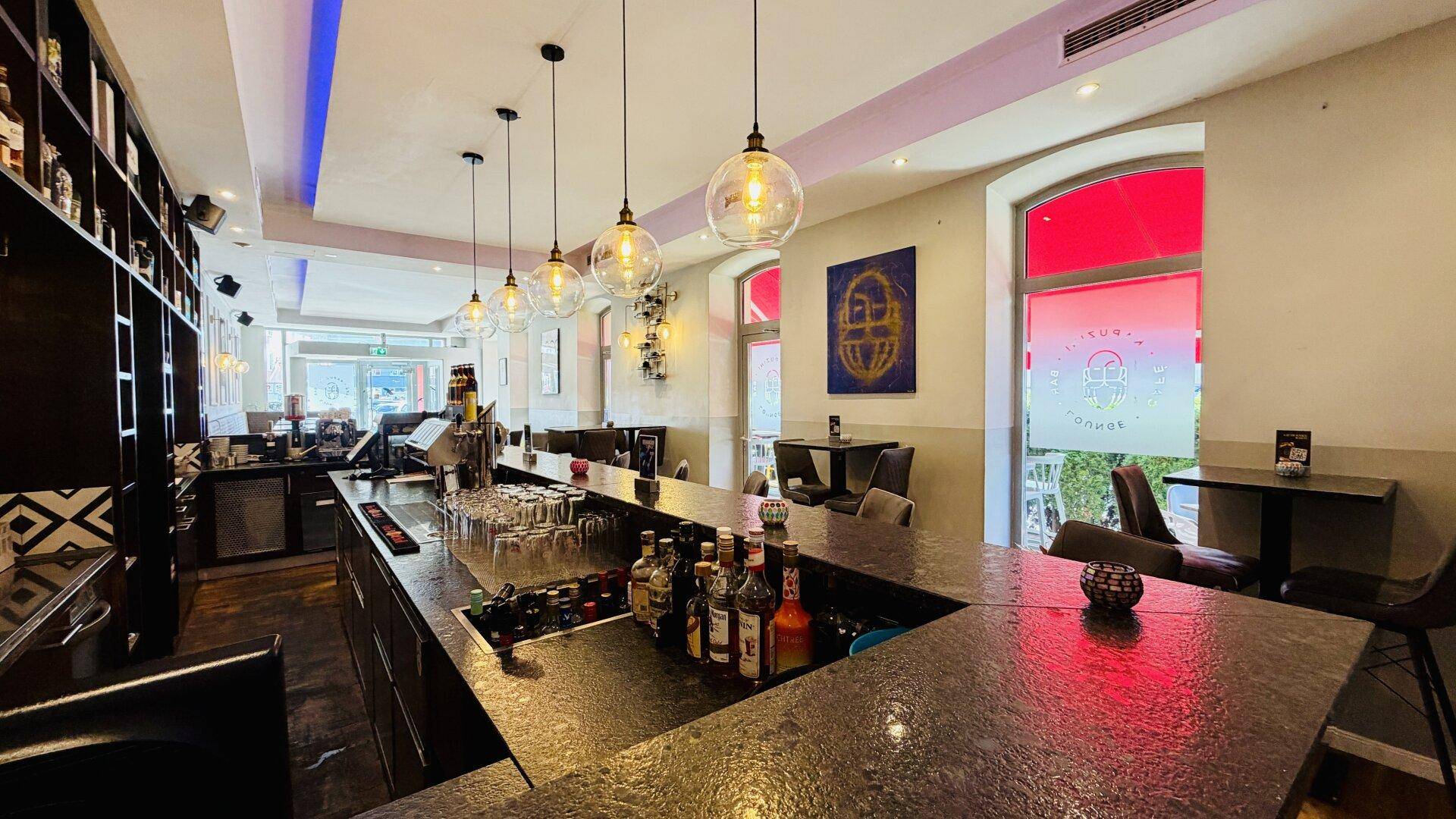 Eine moderne Bar mit dunkler Theke, dekorativen Hängelampen und Blick auf die Außenterrasse.