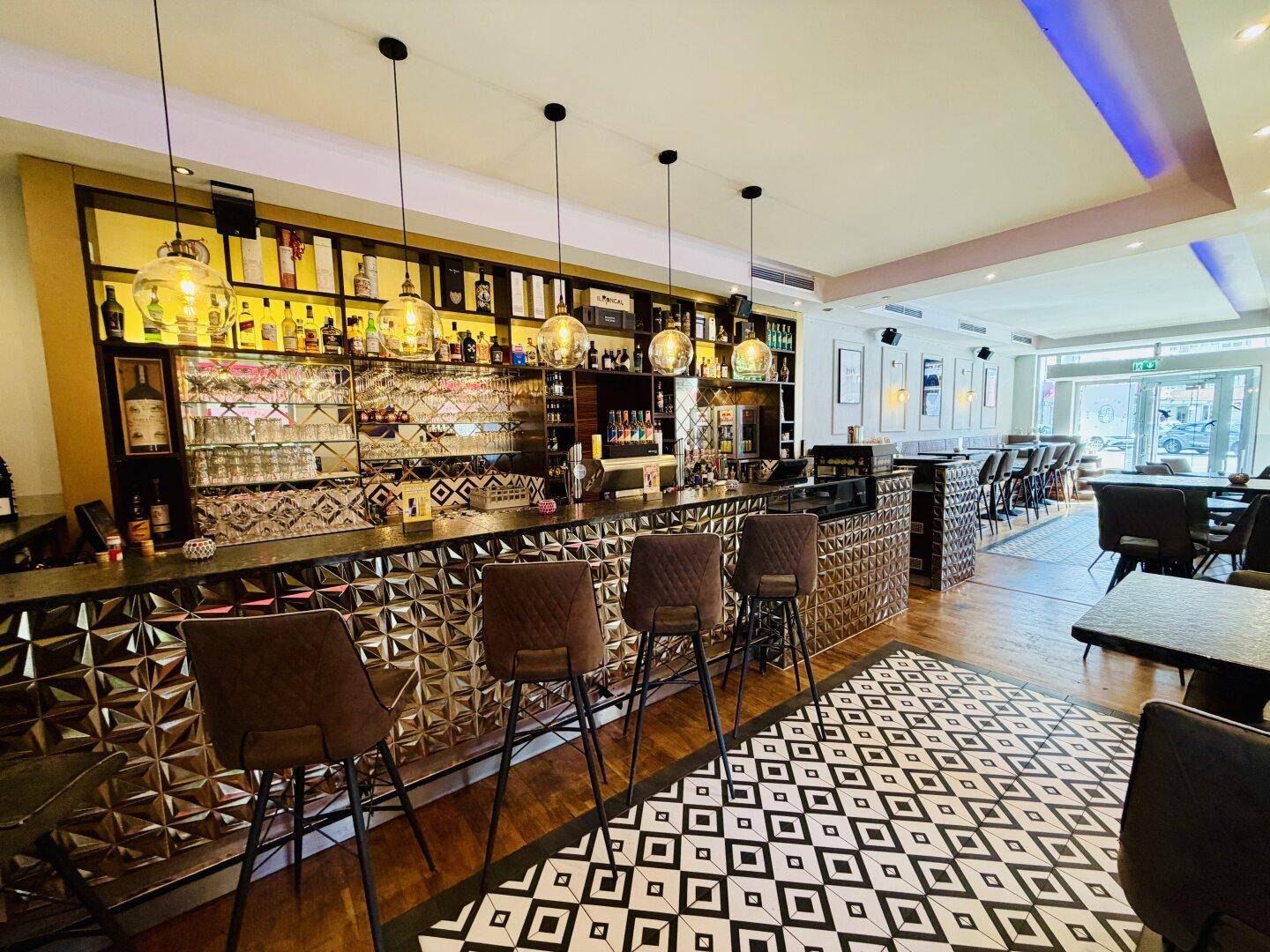 Eine moderne Bar mit dunkler Theke, dekorativen Hängelampen und einem auffälligen gemusterten Boden.